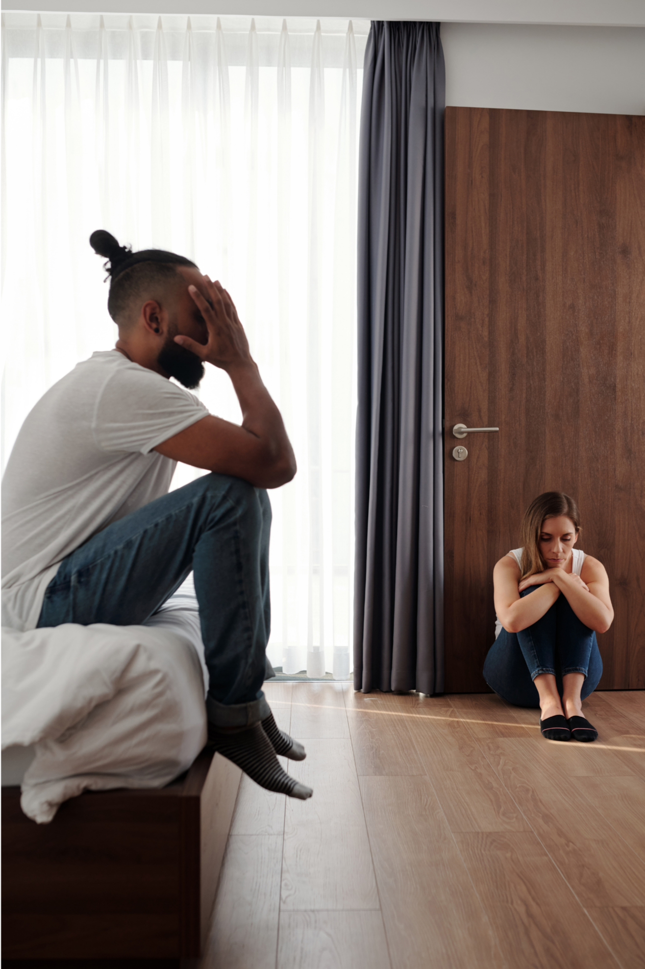 A man sitting on a bed holding his head in distress, a woman sitting on the floor with her arms crossed and head down, appearing upset in a bedroom with window curtains and a wooden door.