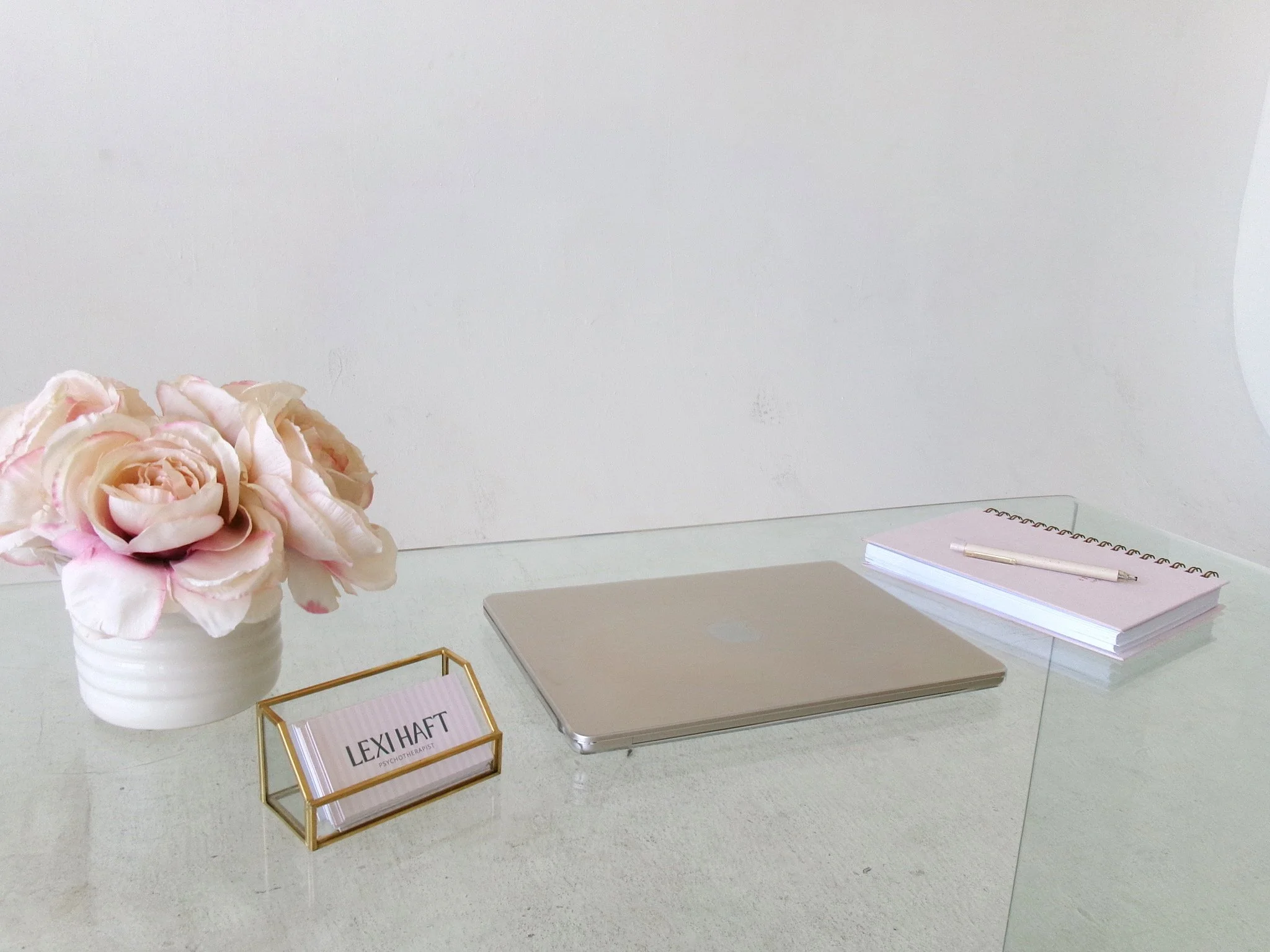 A white desk with a bouquet of pink and white roses in a white vase, a gold desk nameplate reading 'LEXI HAF T', a closed silver laptop, a white notepad with a pen, all set against a plain white wall.