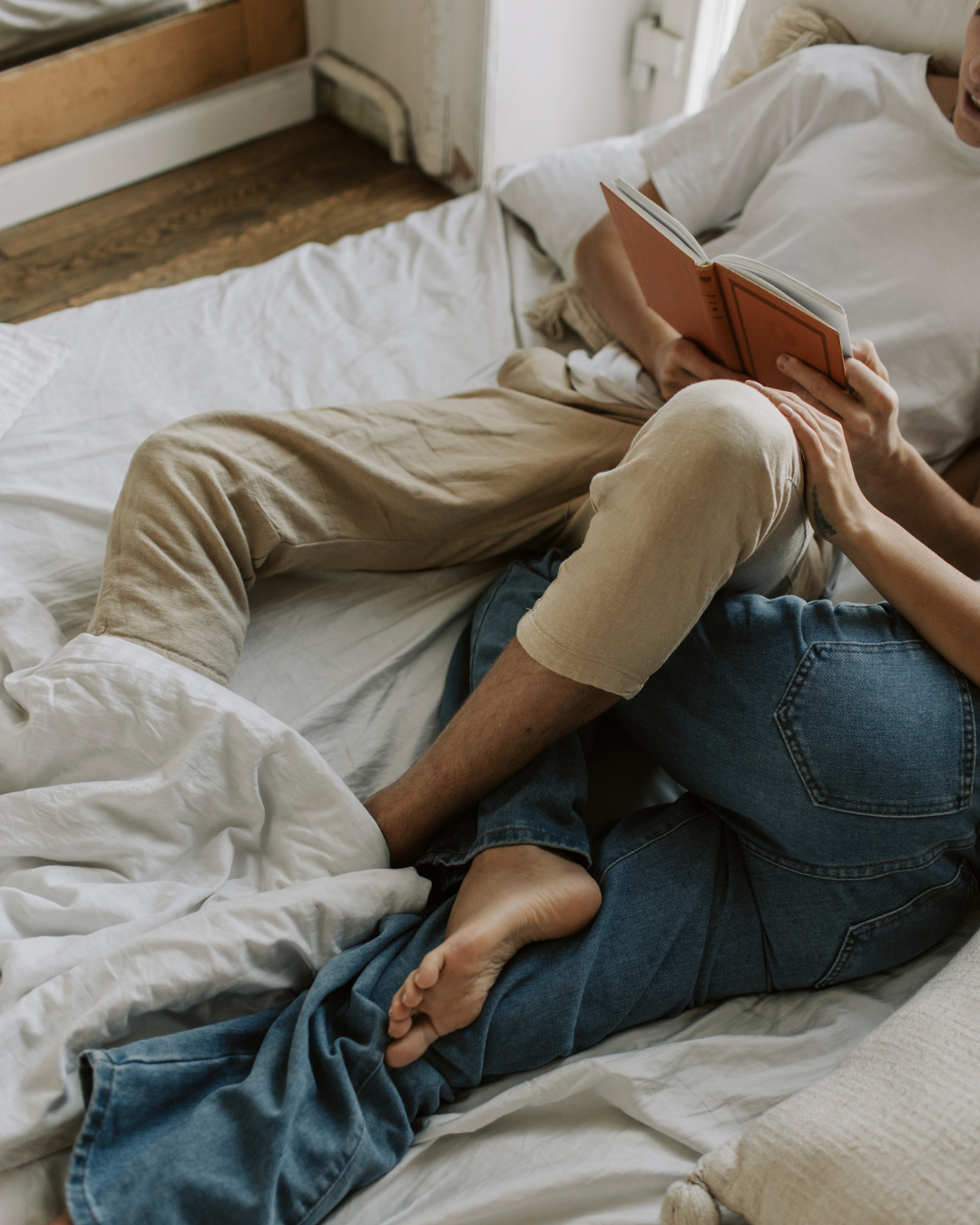 Two people lying on a bed; one reading a book, with their legs intertwined.