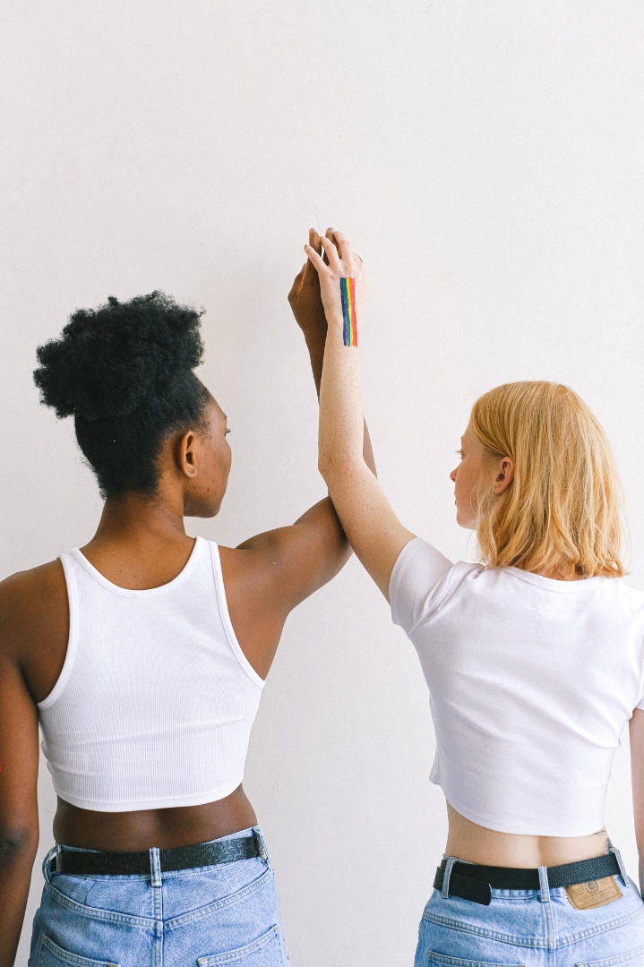 Two women, one with dark curly hair and the other with blonde hair, stand against a plain white wall, touching hands with a rainbow-colored bracelet.