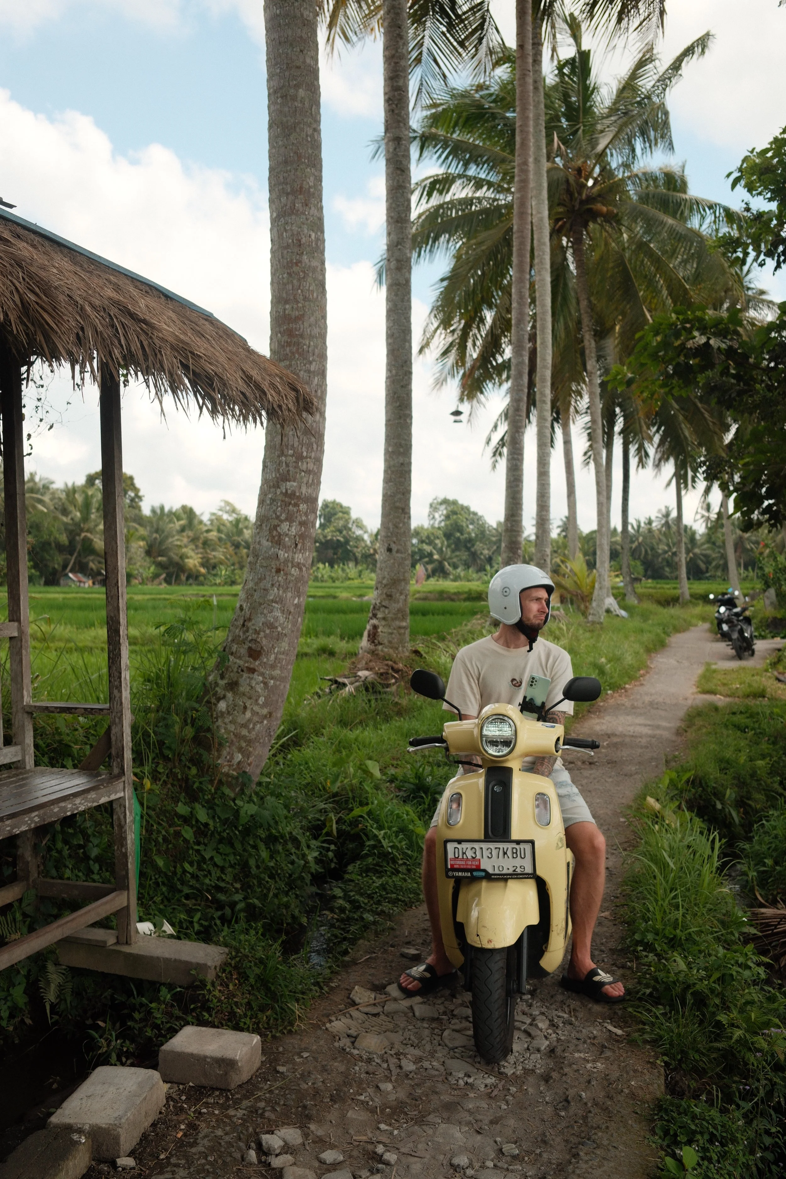 A man wearing a helmet is sitting on a yellow scooter on a narrow dirt path in a rural area with green fields and tall palm trees in the background.