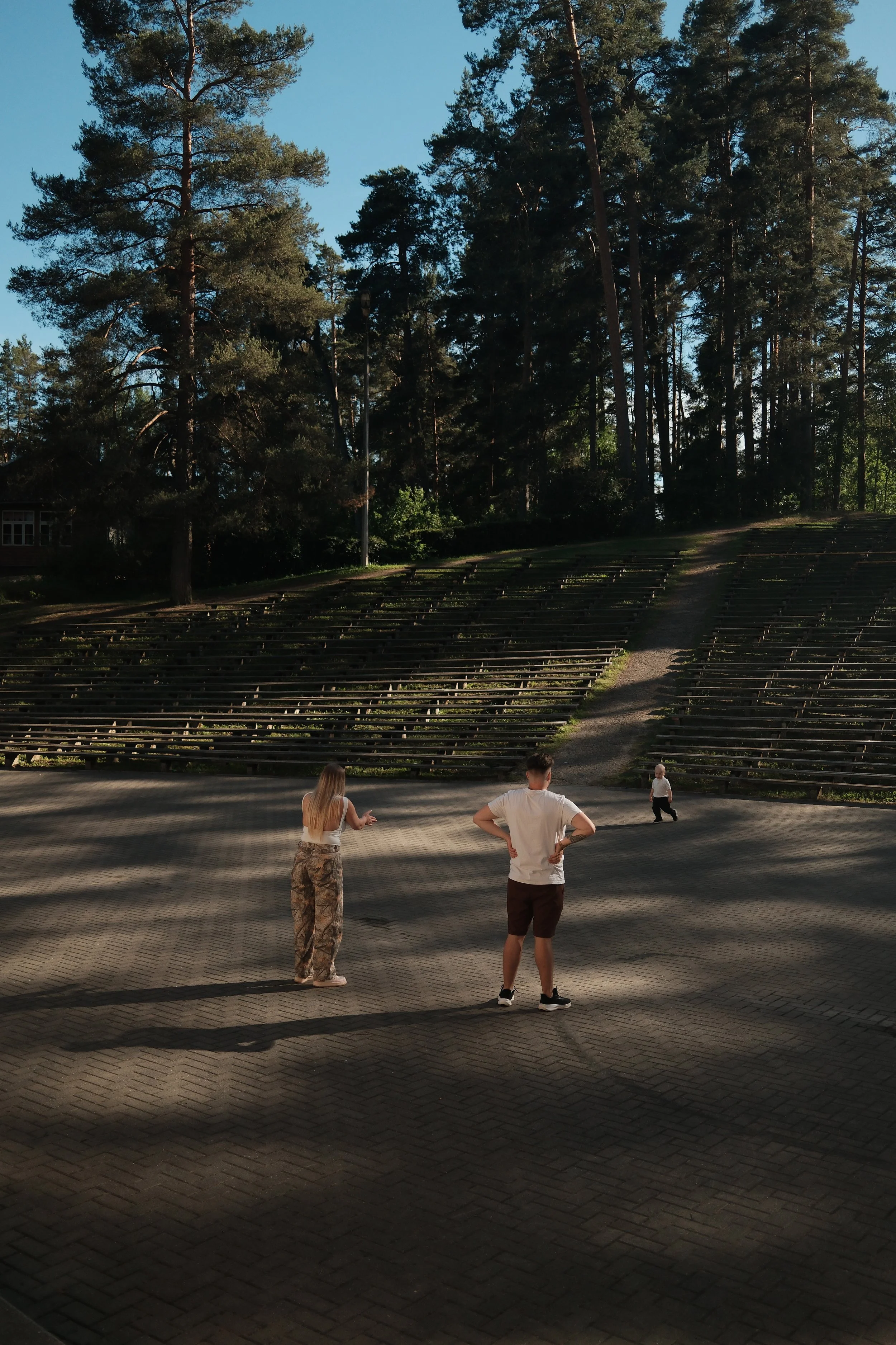 Three people with light skin, a woman and two men, standing on a paved outdoor area, looking towards a forested hill with tiered seating and tall trees, in sunshine with shadows on the ground.