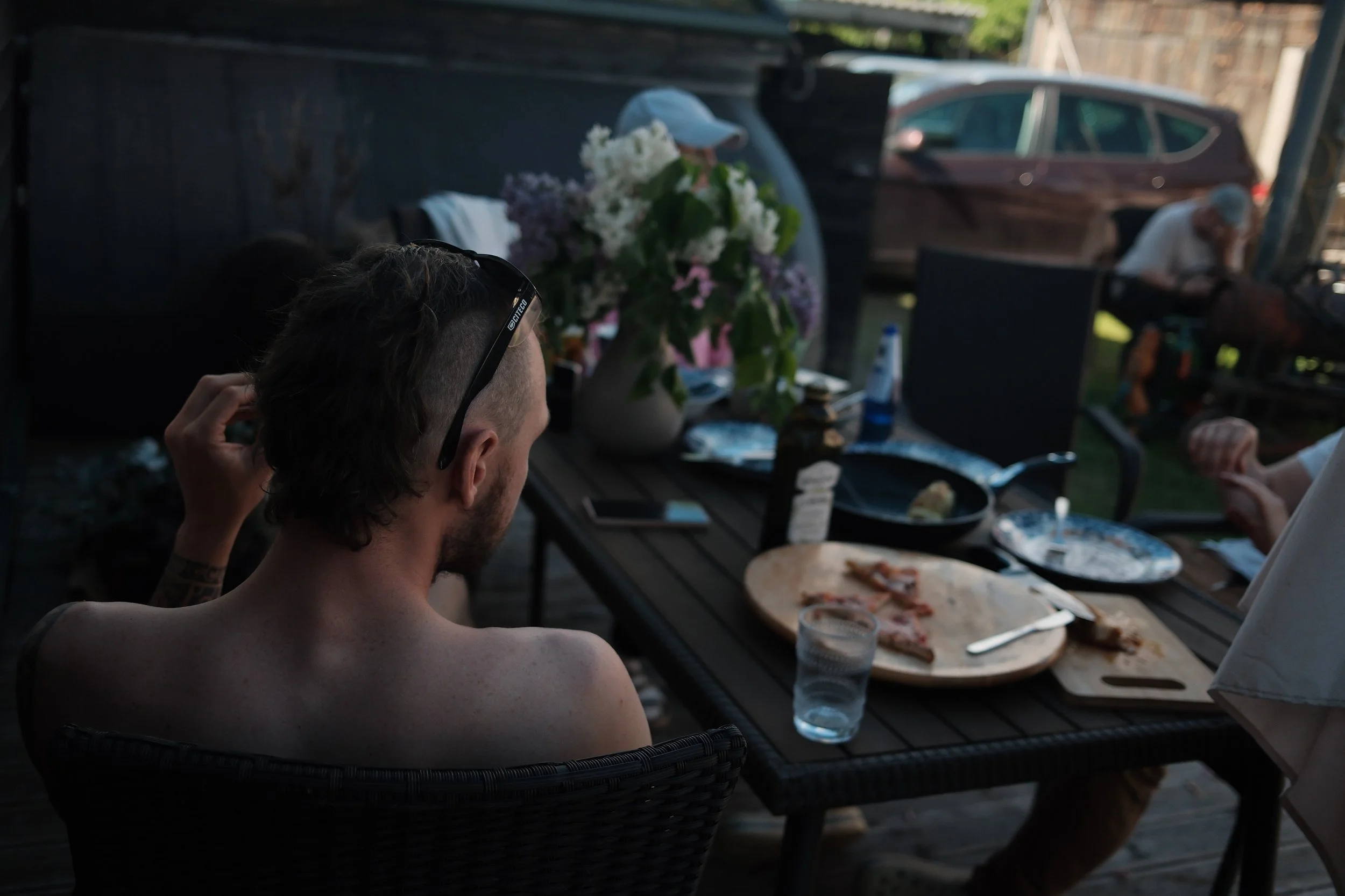 Person with short hair and glasses sitting at an outdoor dining table during sunset with pizza, drinks, and flowers.