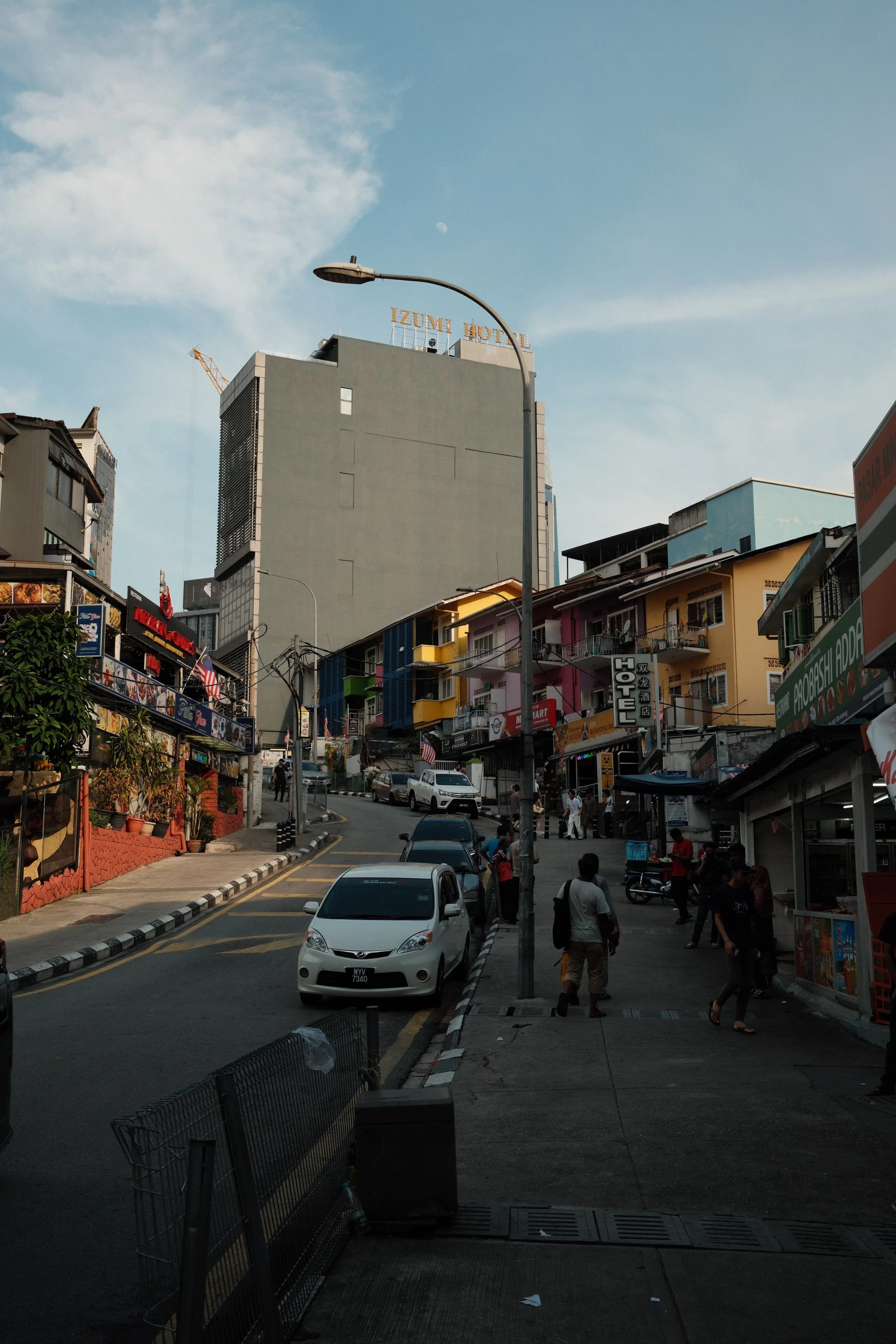 City street scene with cars parked along the curb, a sidewalk with pedestrians, colorful buildings, and a tall gray building with a sign that reads "IZUMI HOTEL" in the background.