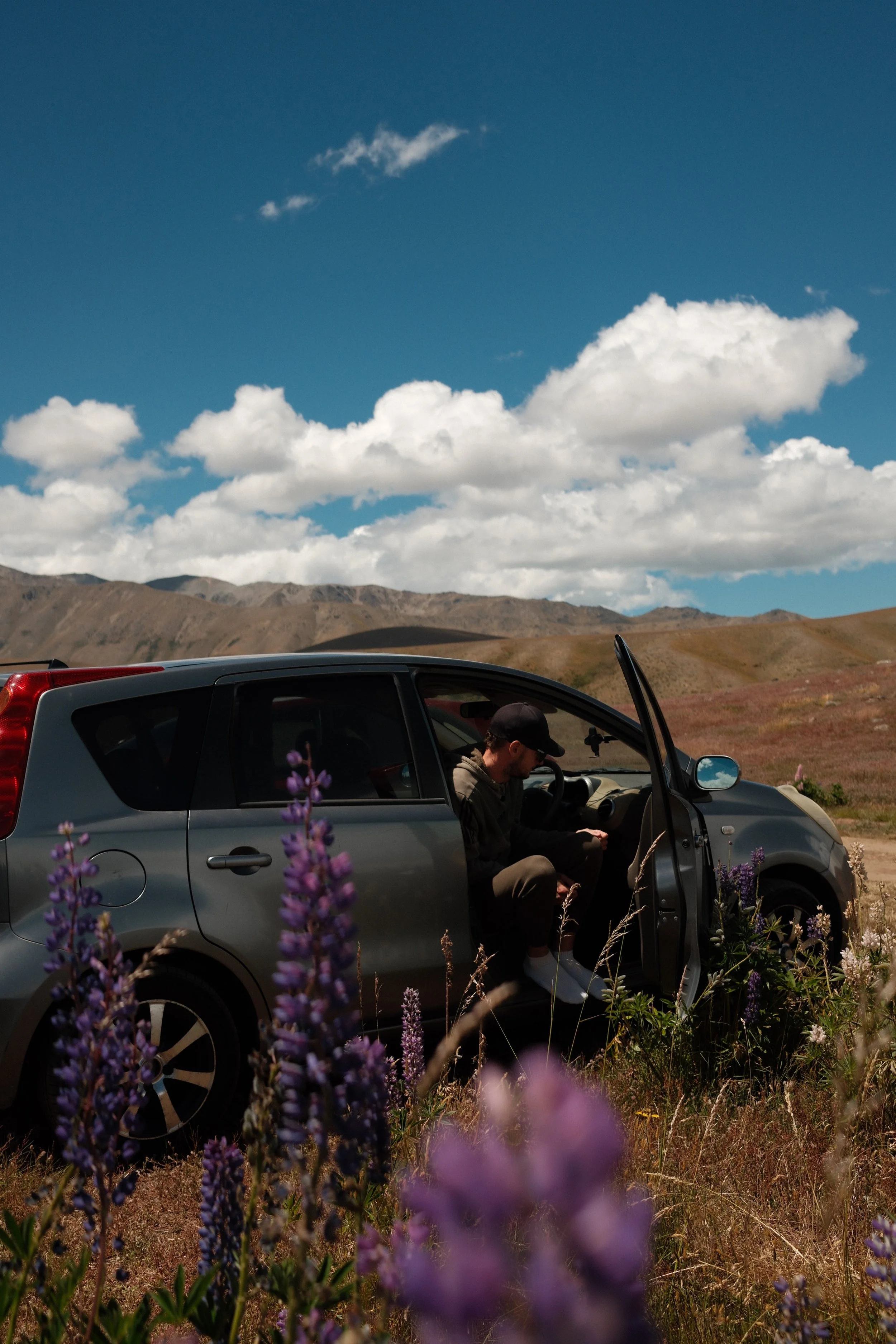 A man sitting in a parked car on a rural landscape with mountains and a partly cloudy sky, surrounded by purple flowers in the foreground.