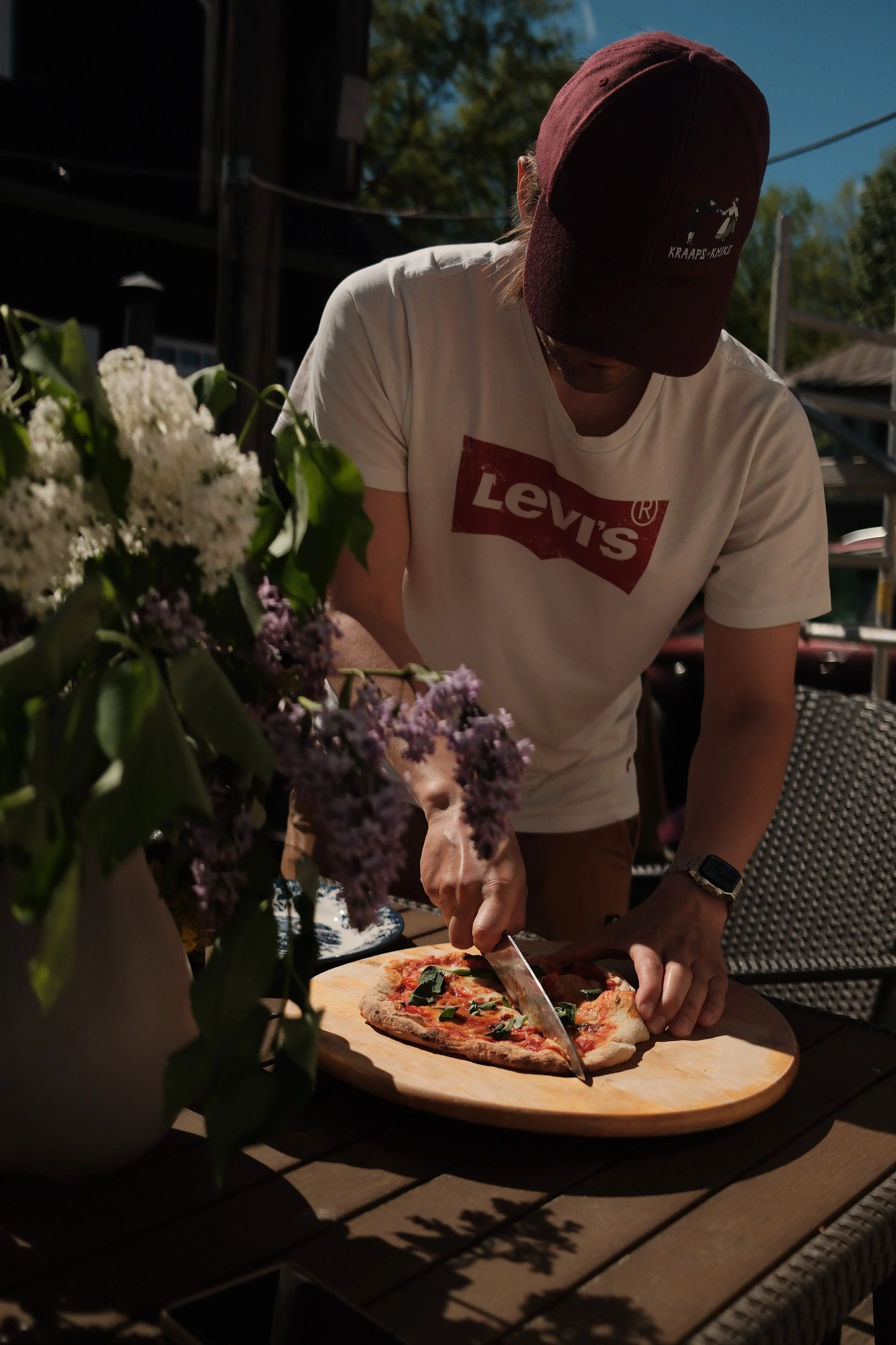 A person wearing a white Levi's T-shirt and a maroon cap is slicing a pizza on a wooden cutting board outdoors. There are flowers in the foreground and a structure with trees in the background.