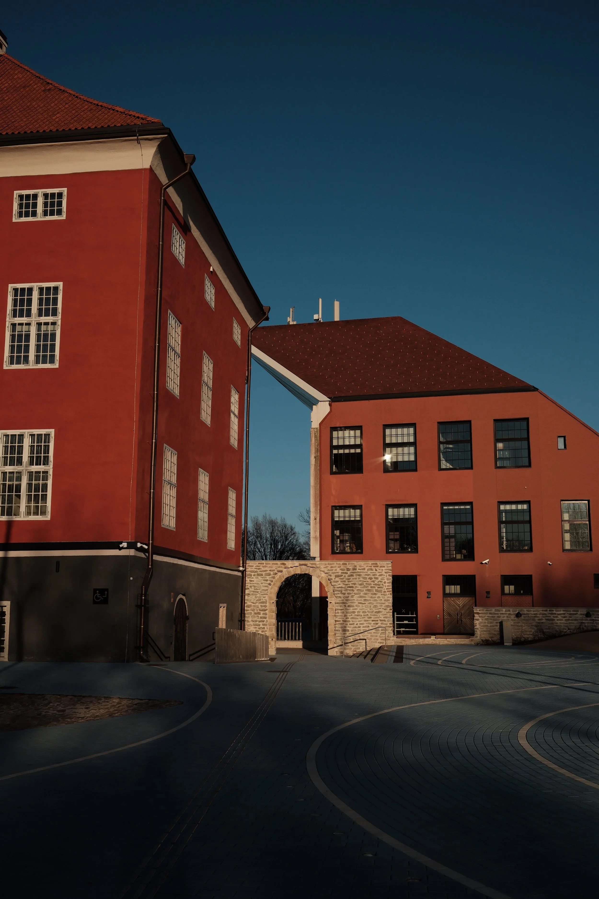 Two modern buildings with red and orange facades under a clear blue sky, featuring large windows. An arched stone gateway connects parts of the buildings, with a paved courtyard and curved street in the foreground.