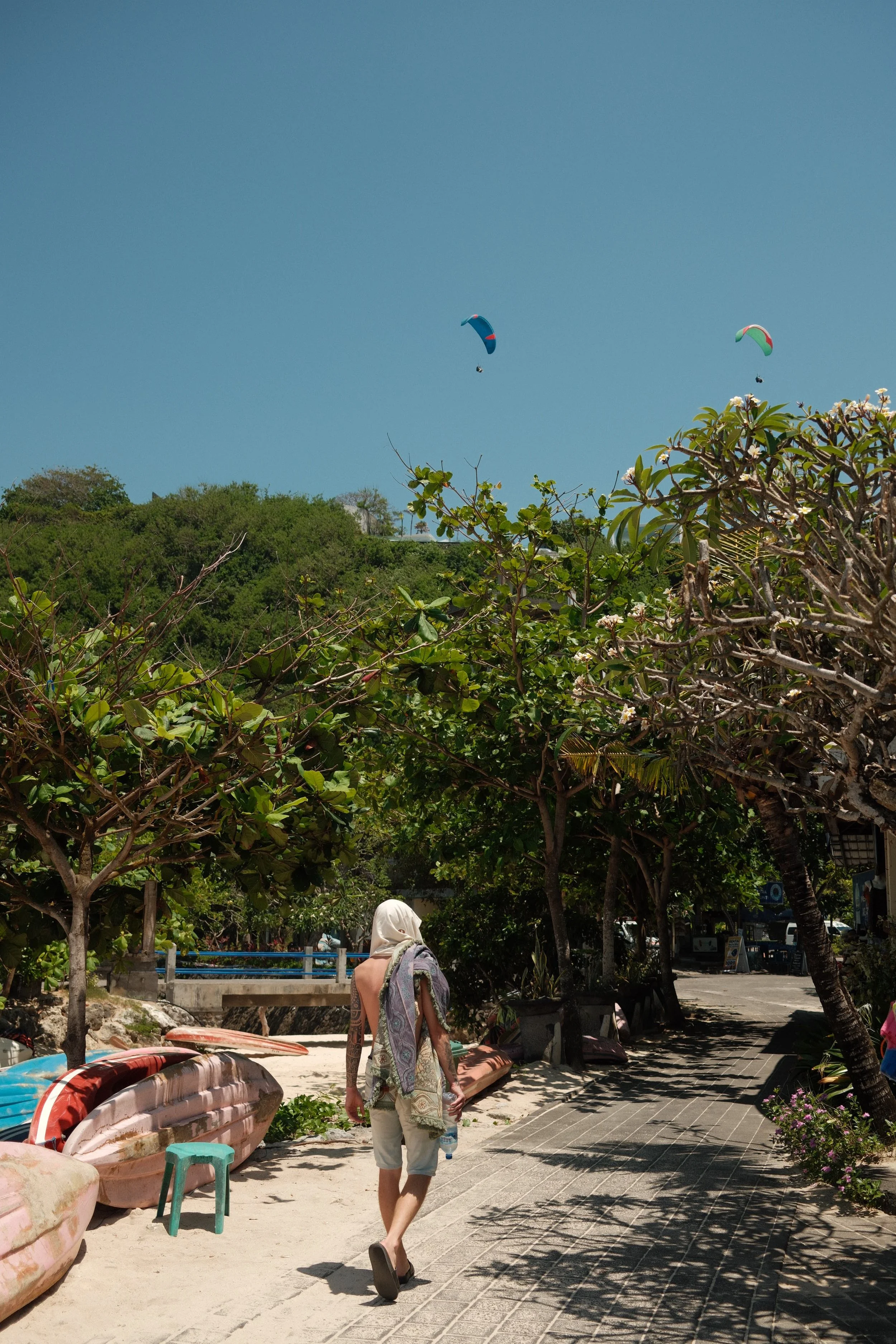 Woman walking along a sandy path with boats and lush green trees on a sunny day, with two paragliders flying in the clear blue sky.