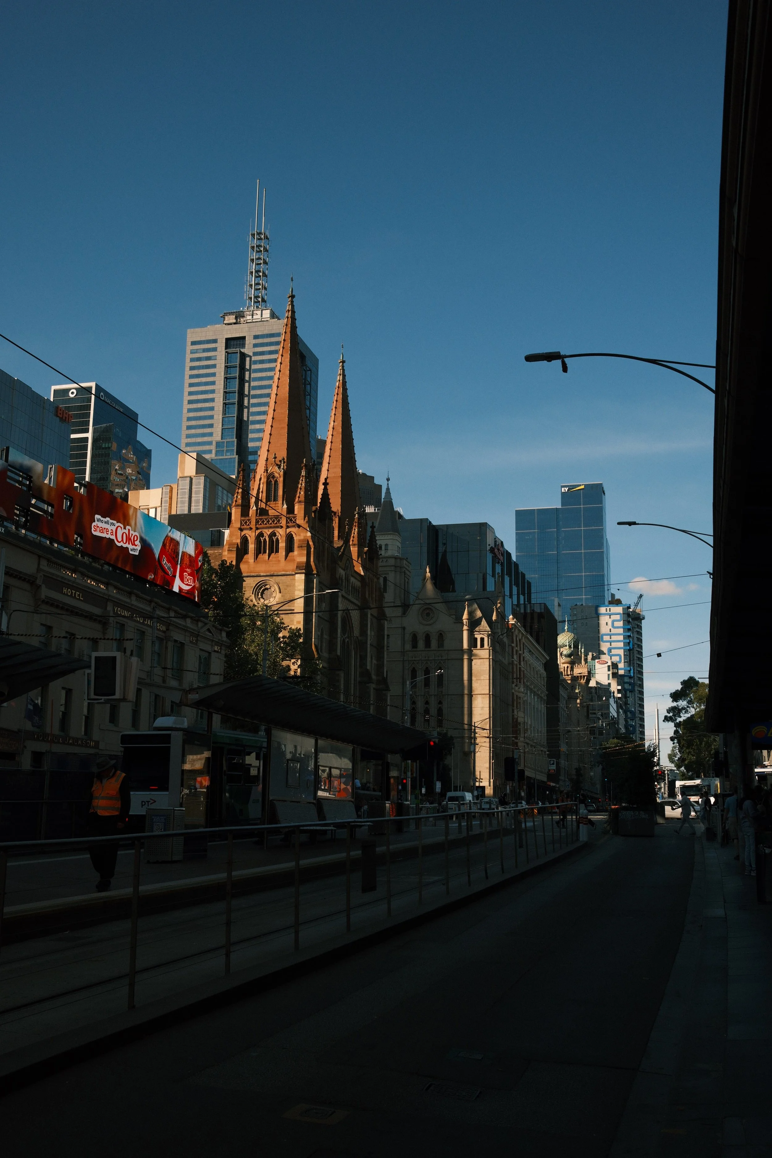 A city street scene with a mix of modern and historic architecture, including a church with pointed spires, tall skyscrapers, a blue sky, and a tram stop with a few pedestrians and a tram in the foreground.