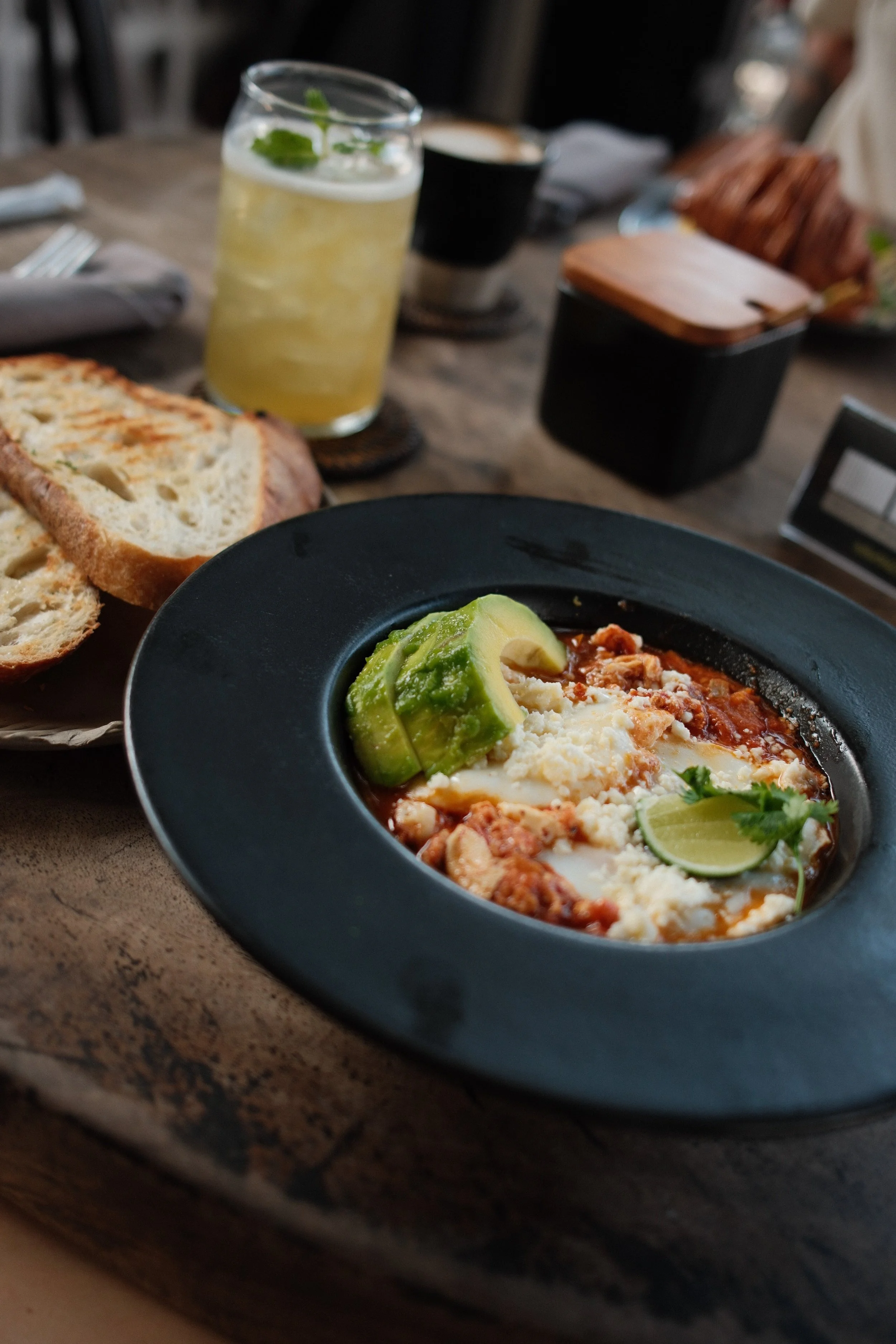 A black bowl of Mexican-style breakfast food with cheese, eggs, avocado slices, and cilantro, on a rustic wooden table alongside slices of toasted bread, a glass of limeade with mint, and a cup of coffee.