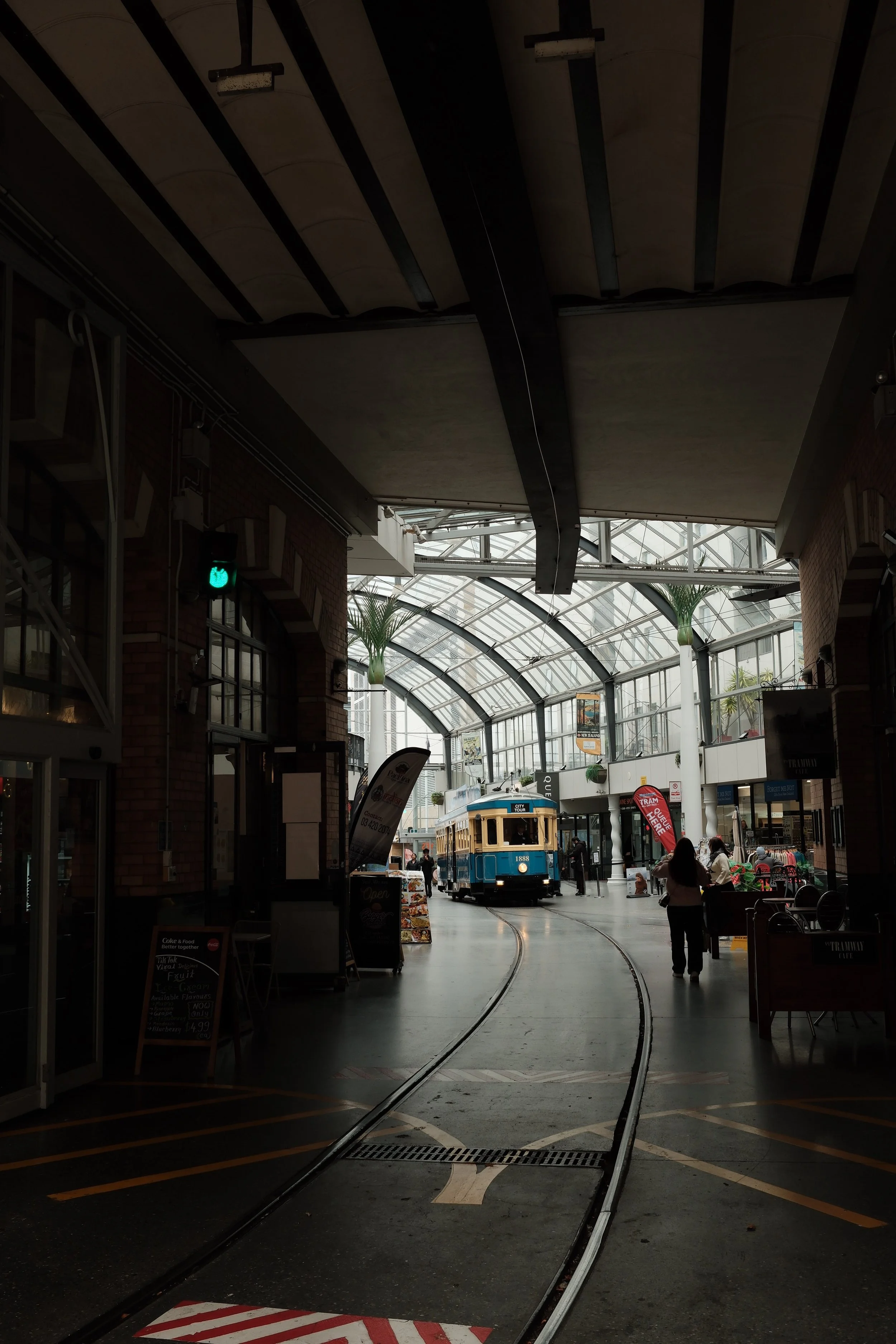 Indoor shopping mall with tram tracks and a vintage blue tram in the center. The mall has a high glass ceiling with natural light, decorative plants, and storefronts on the sides with people walking and shopping.