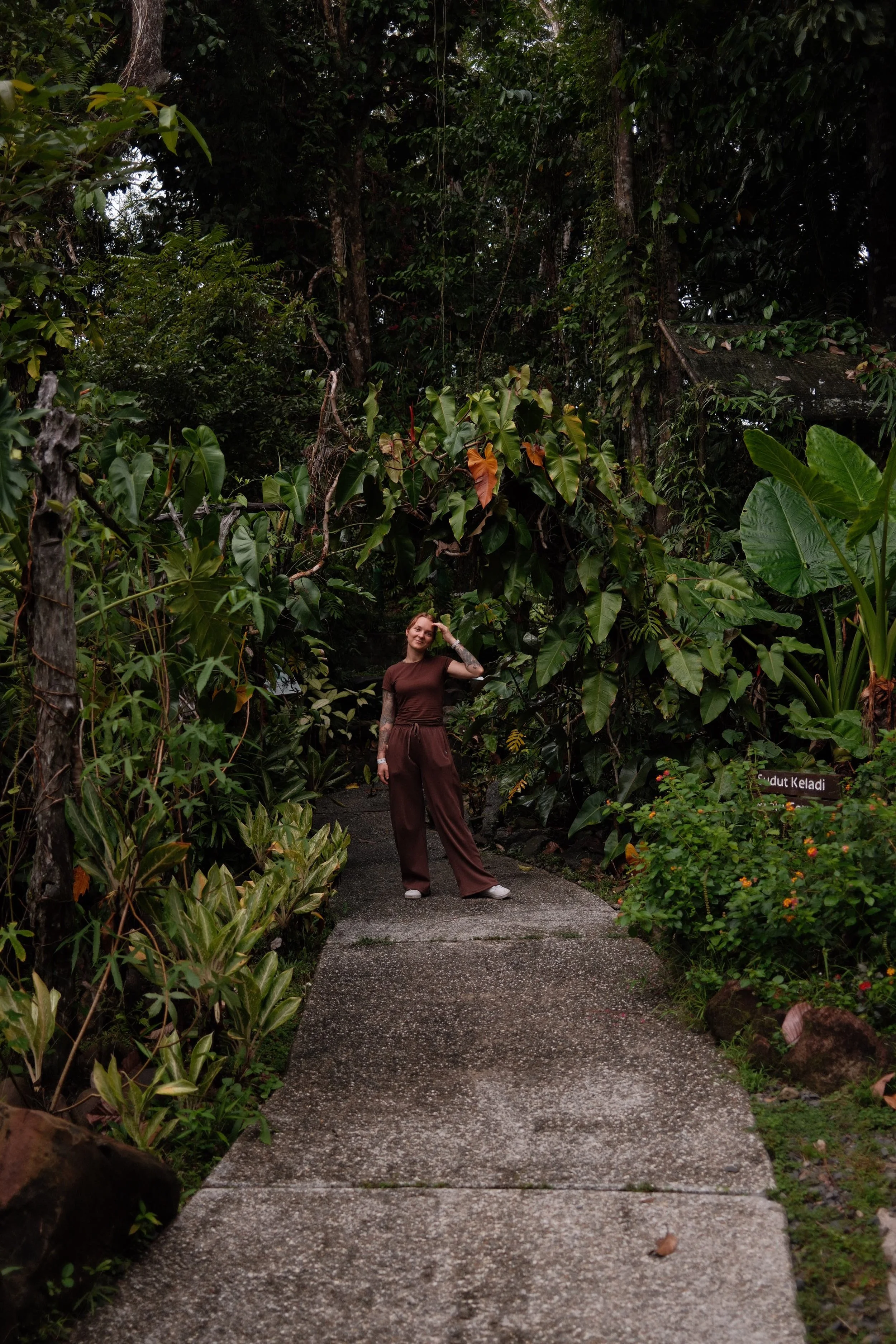 A woman with tattoos wearing a brown outfit standing on a lush, green jungle trail, surrounded by dense tropical plants and trees, with a sign that reads 'Mandut Keladi' on the right side.