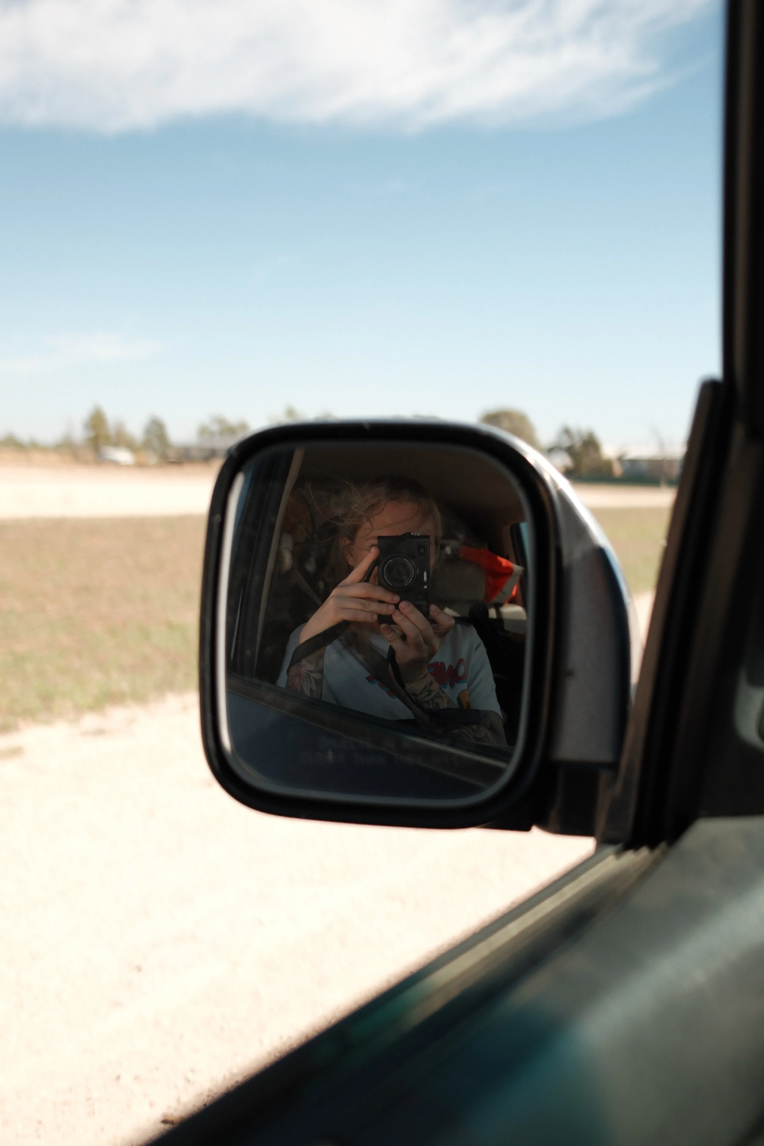 Photo of a person taking a photo of themselves in a car's side mirror with blue sky and some trees in the background.