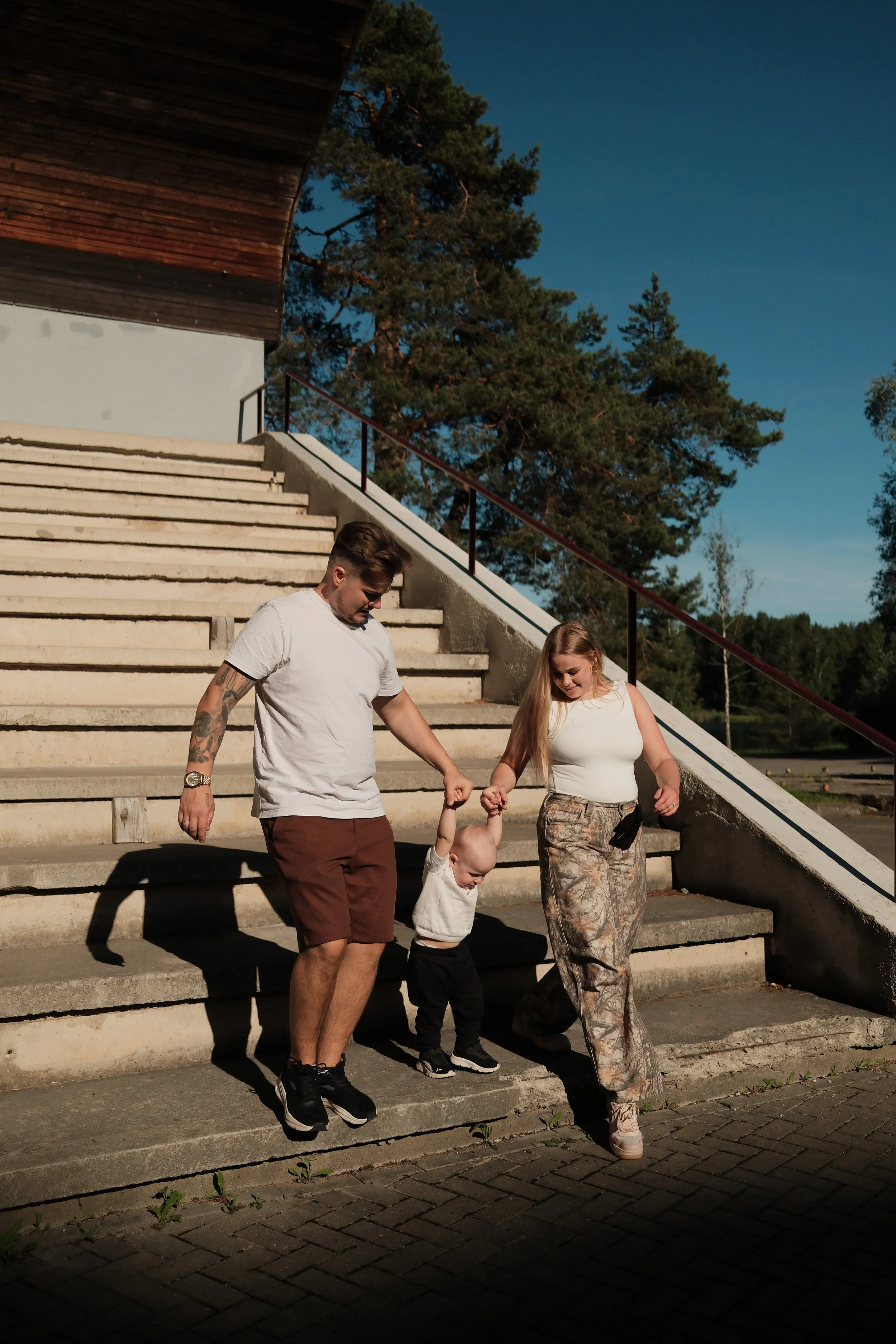 A family of four walking down outdoor concrete stairs during sunset, holding hands, with trees and a clear sky in the background.