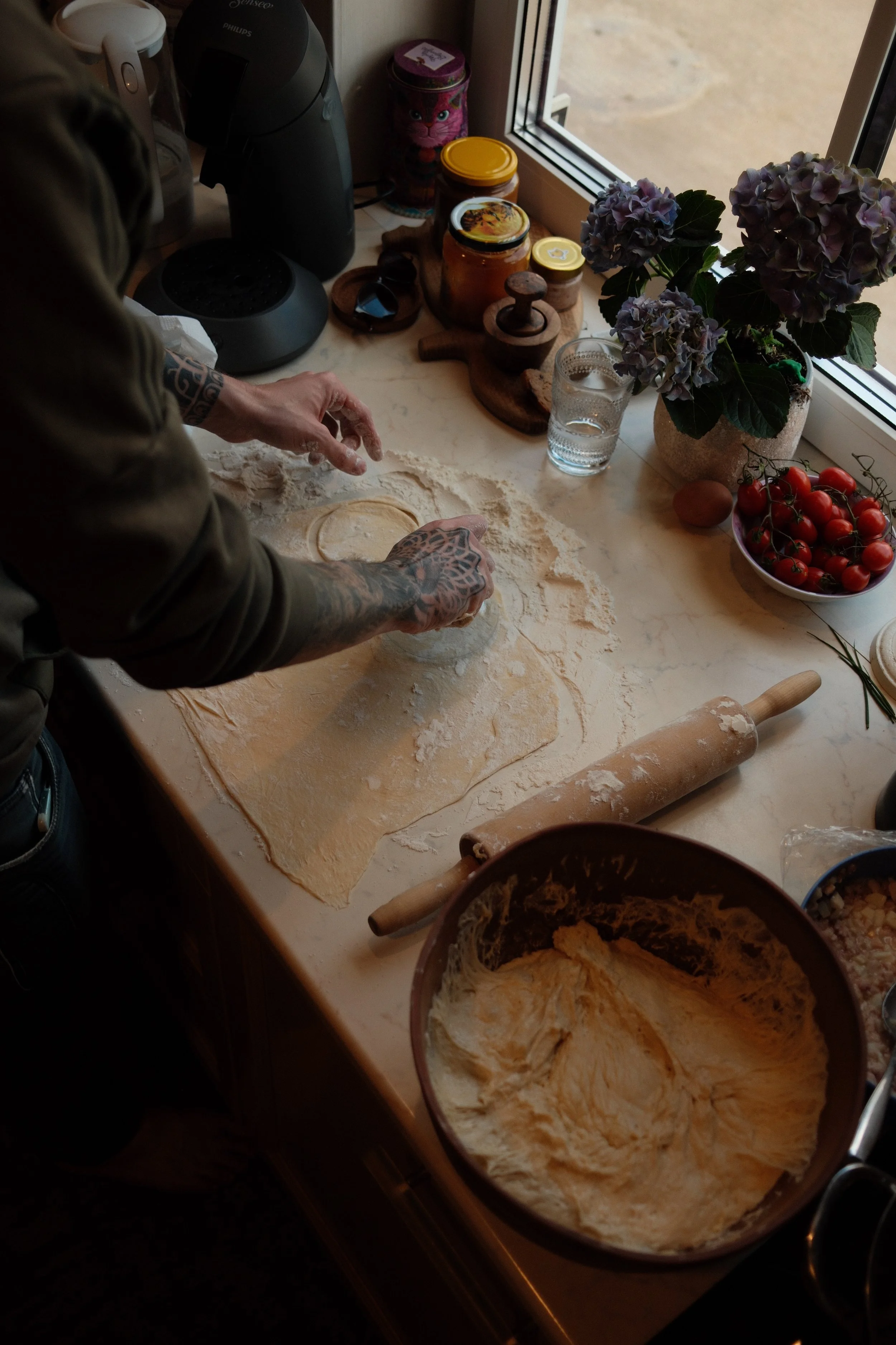 A person preparing dough on a kitchen counter with various ingredients like tomatoes, flowers, honey jars, and kitchen appliances visible around.