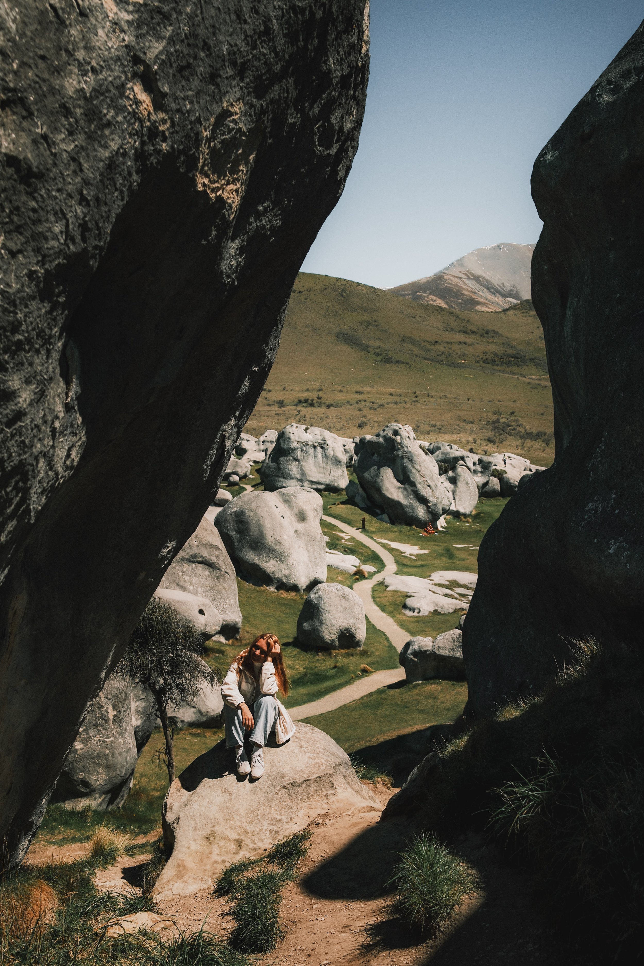 A woman with long hair sitting on a large rock among giant stones in a mountainous landscape, with a trail winding through green grass and hills in the background.
