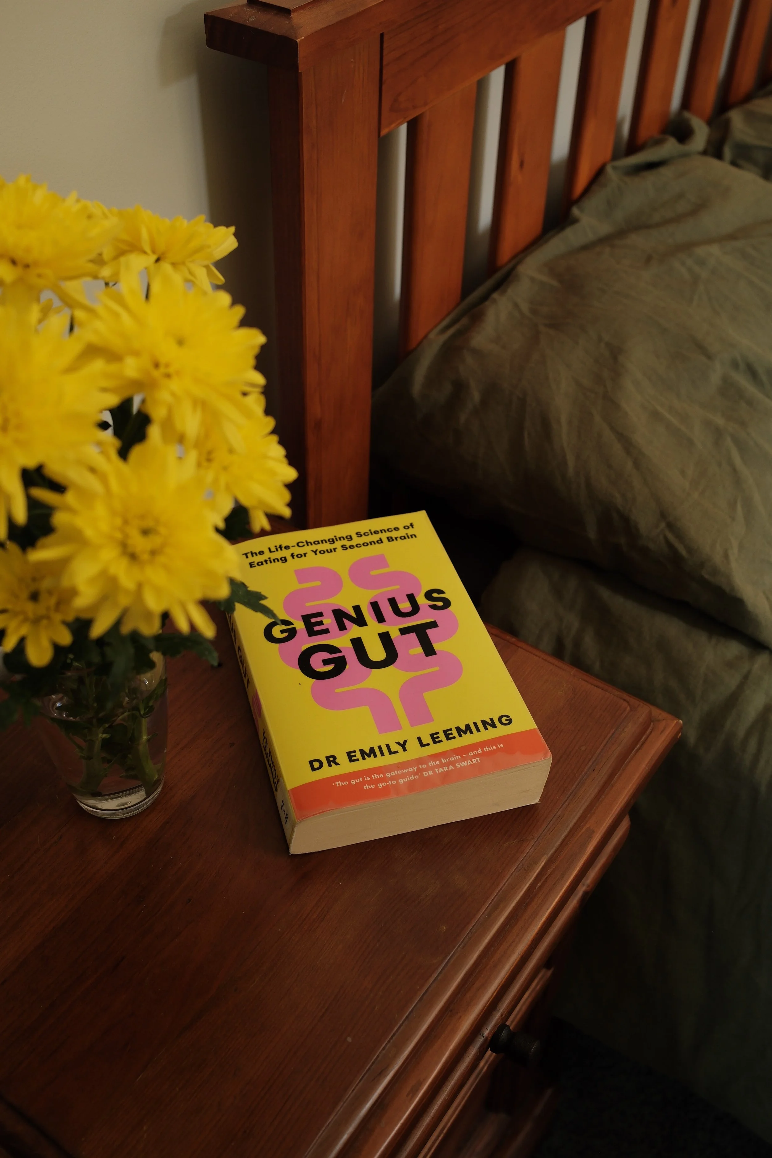 A wooden nightstand with a yellow book titled "Genius Gut" by Dr. Emily Leeming, a glass of yellow flowers, and a bed with brown bedding in a bedroom setting.