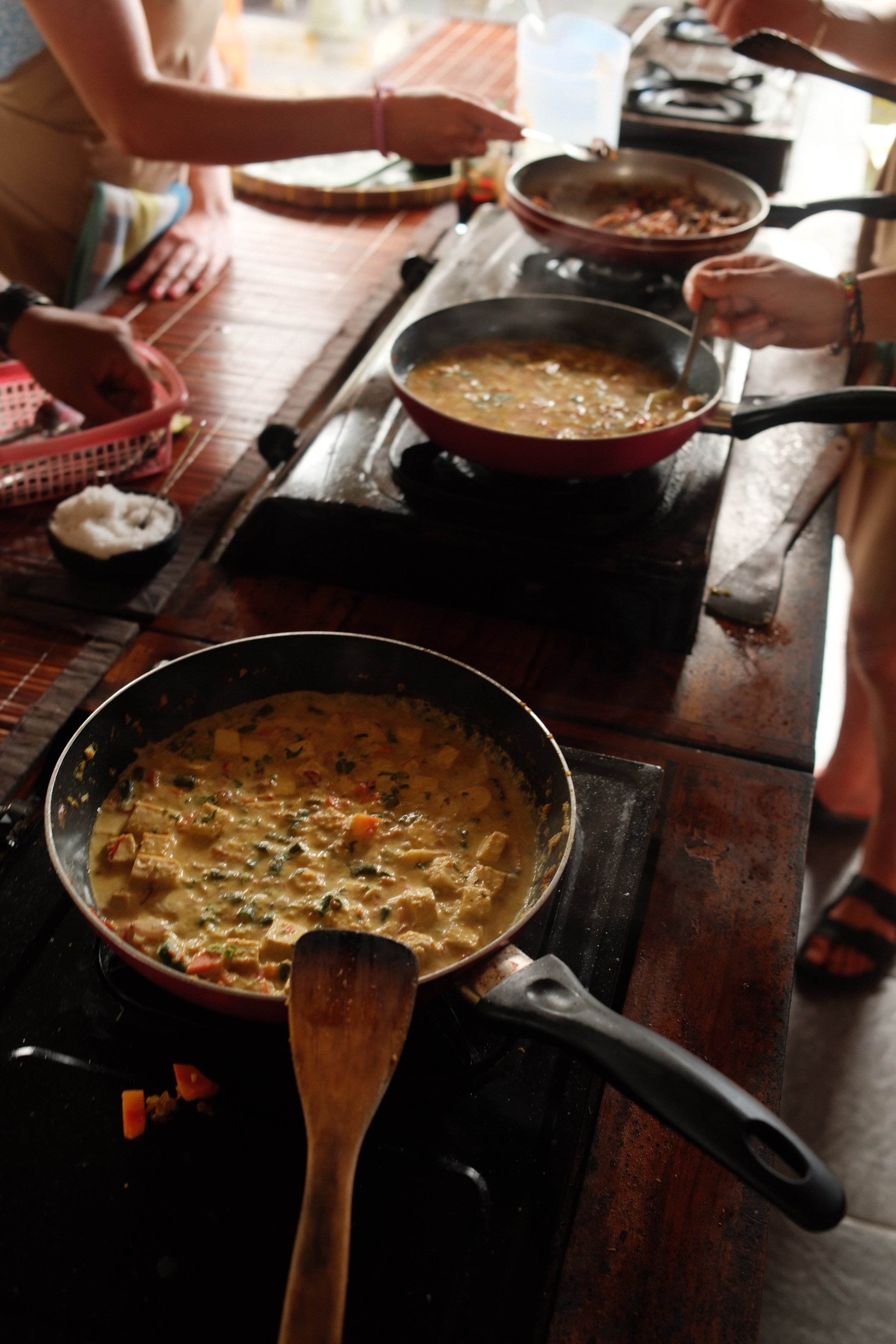 People cooking various dishes on a stove, with a focus on a pan containing a vegetable and tofu stew or curry.