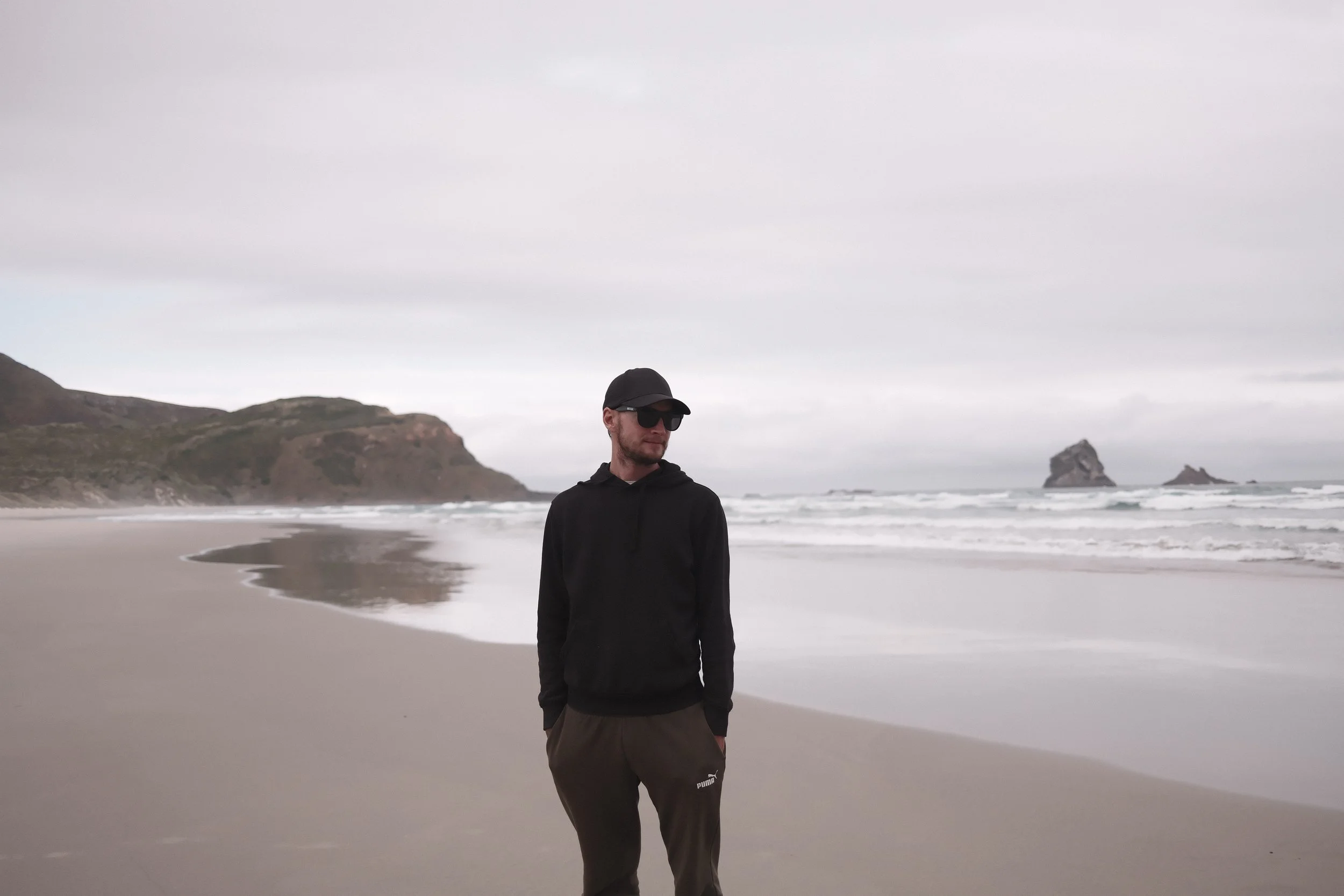A man wearing a black cap, sunglasses, a black hoodie, and gray pants standing on a beach with cliffs and rock formations in the background under an overcast sky