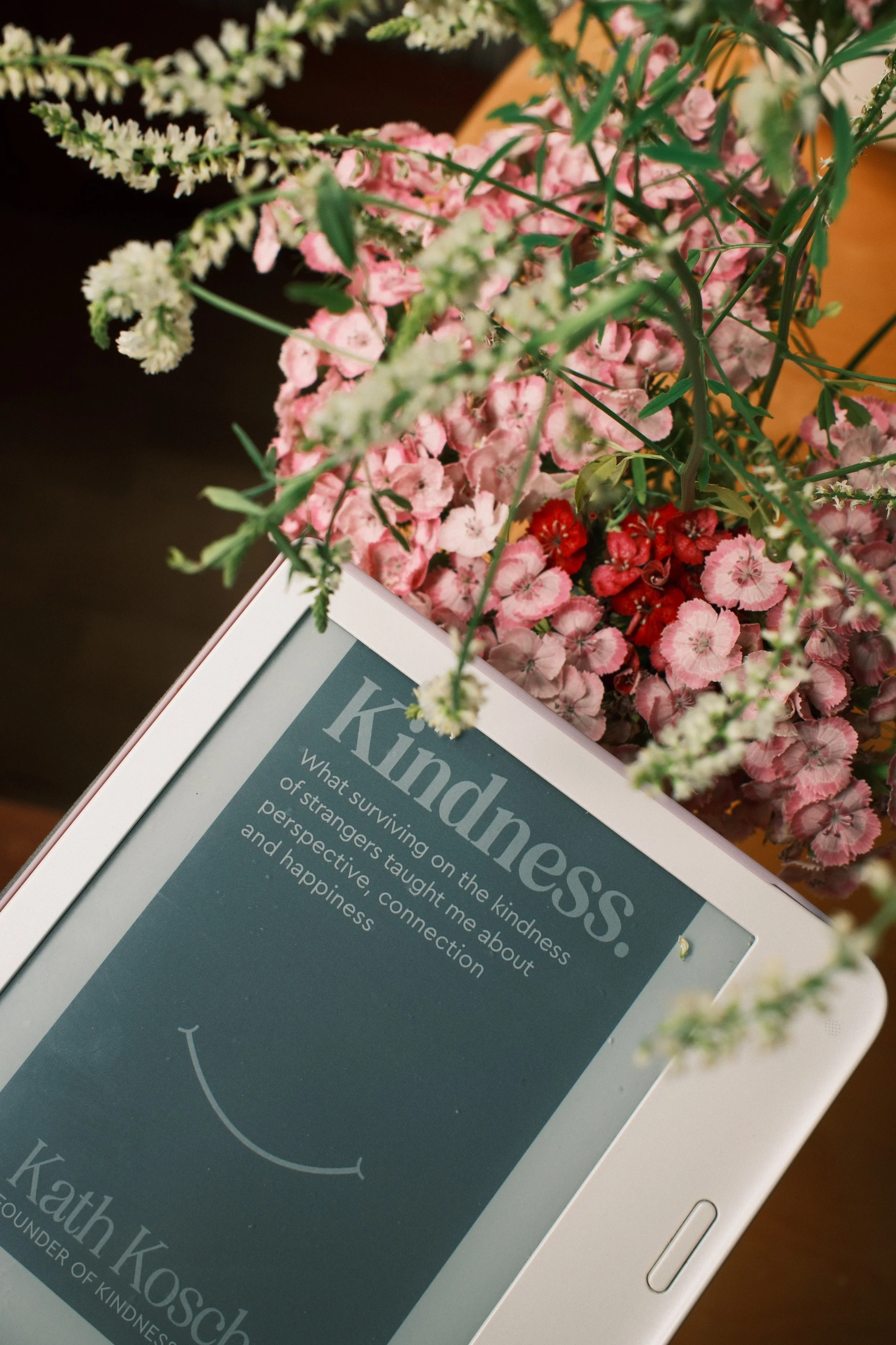 Close-up of a tablet on a table with an article about kindness and happiness, surrounded by pink and white flowers.