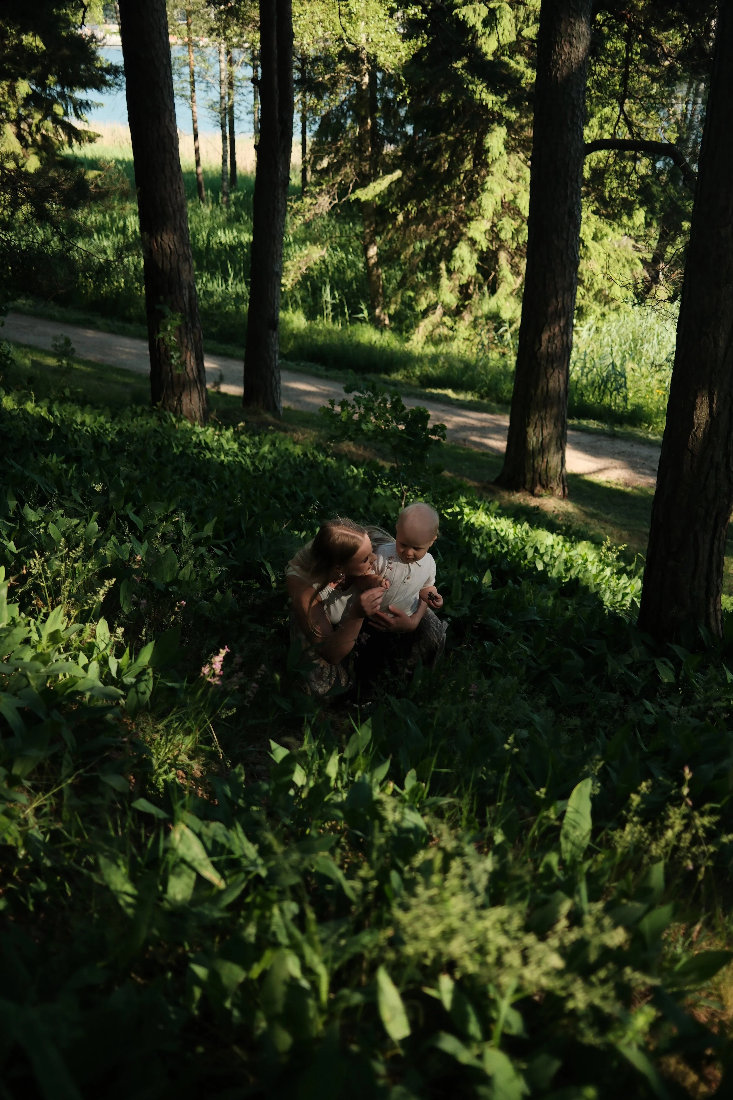 A woman and a young girl are sitting in tall grass and bushes in a wooded outdoor area with tall trees, green foliage, and a dirt path visible, enjoying a moment together.