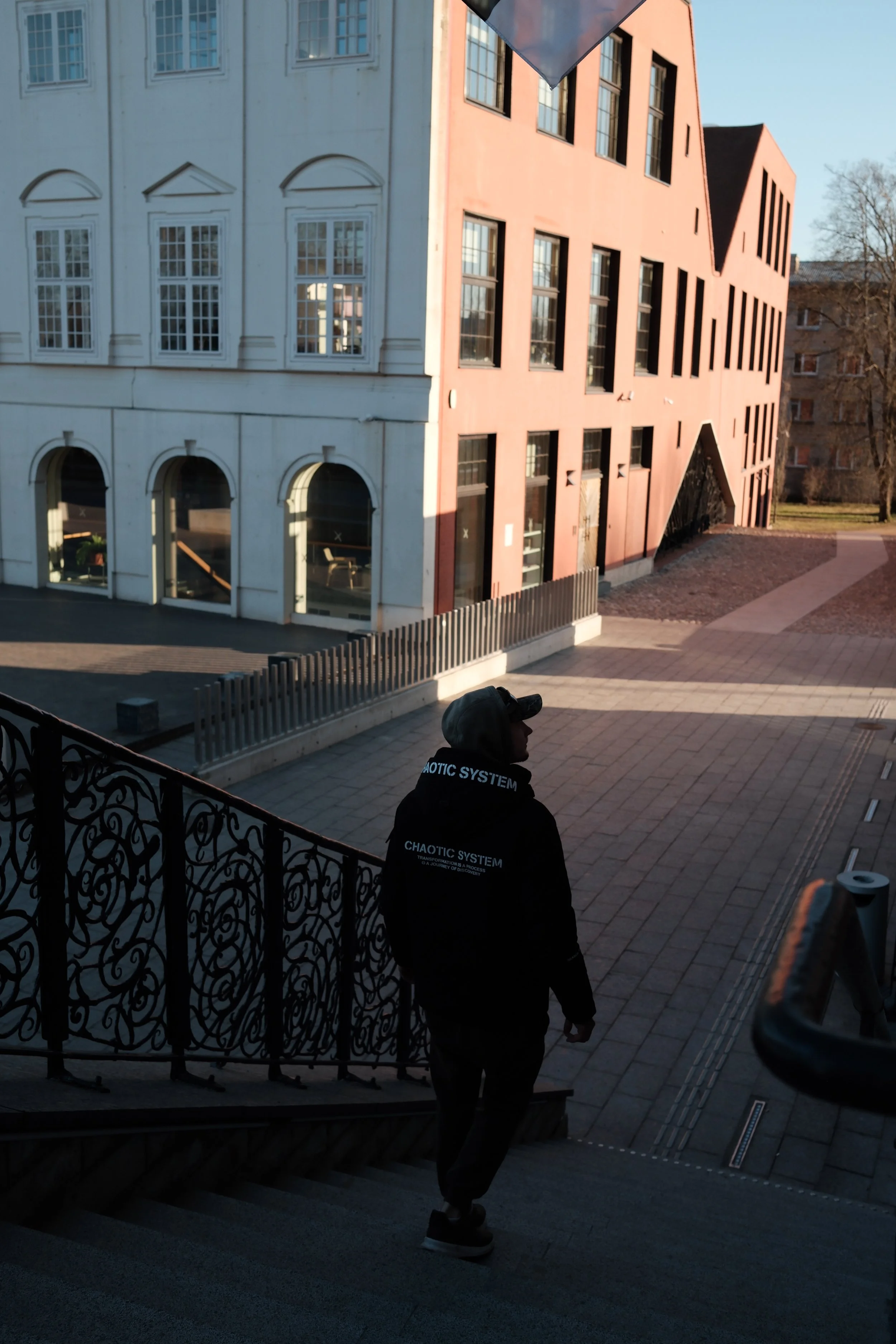 Person wearing a black hoodie with white writing walking down stairs outside a building with architectural design on the facade, sunlight casting shadows.