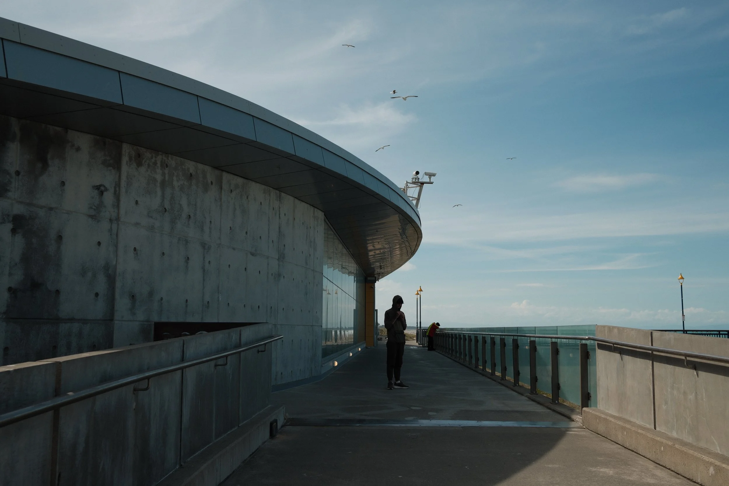 People standing along a coastal promenade next to a modern building with a curved roof and large glass windows, seagulls flying overhead, cloudy sky, and a lamppost in the distance.