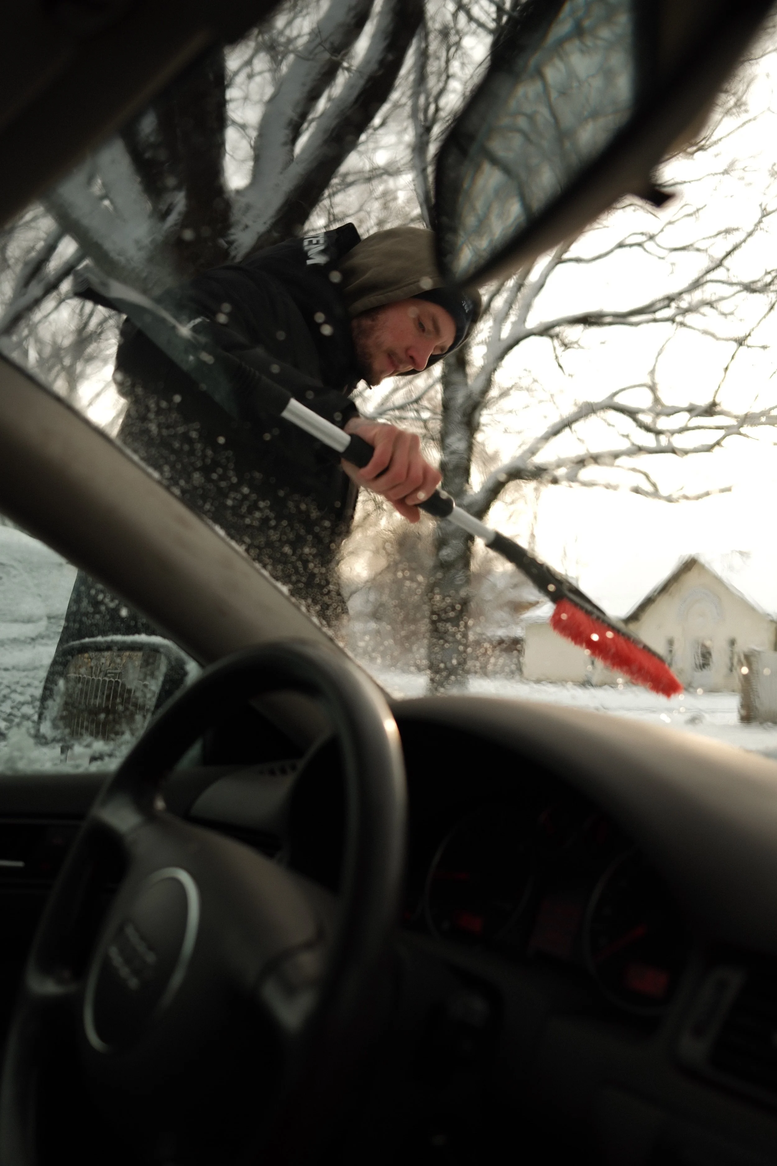 A person cleaning snow off a car's windshield from the inside, seen from the driver's seat with a view through the window showing snow-covered trees and outdoor area.