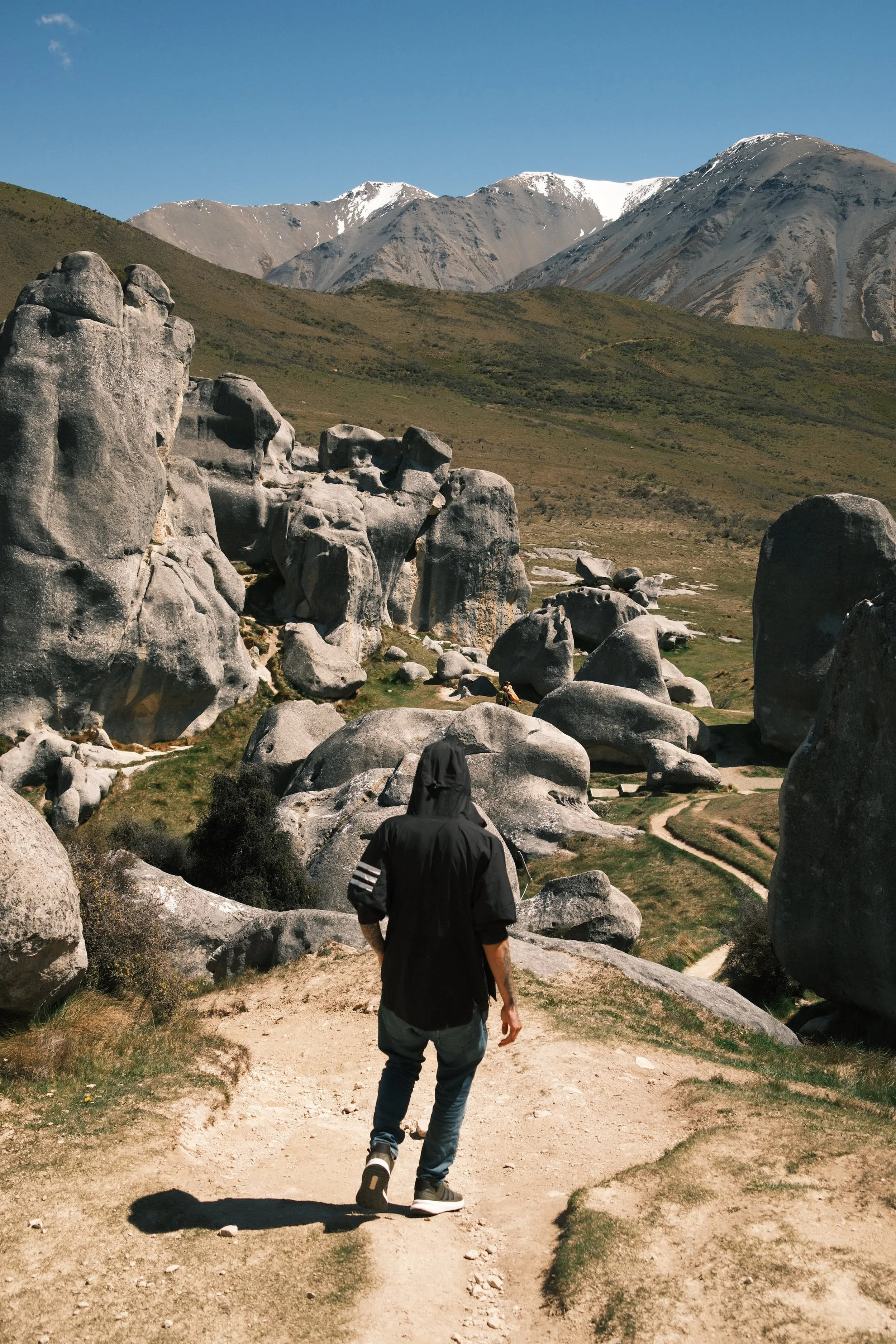 A person wearing a black hoodie and jeans walking on a dirt path surrounded by large rocks and boulders, with a mountain range in the background under a clear blue sky.