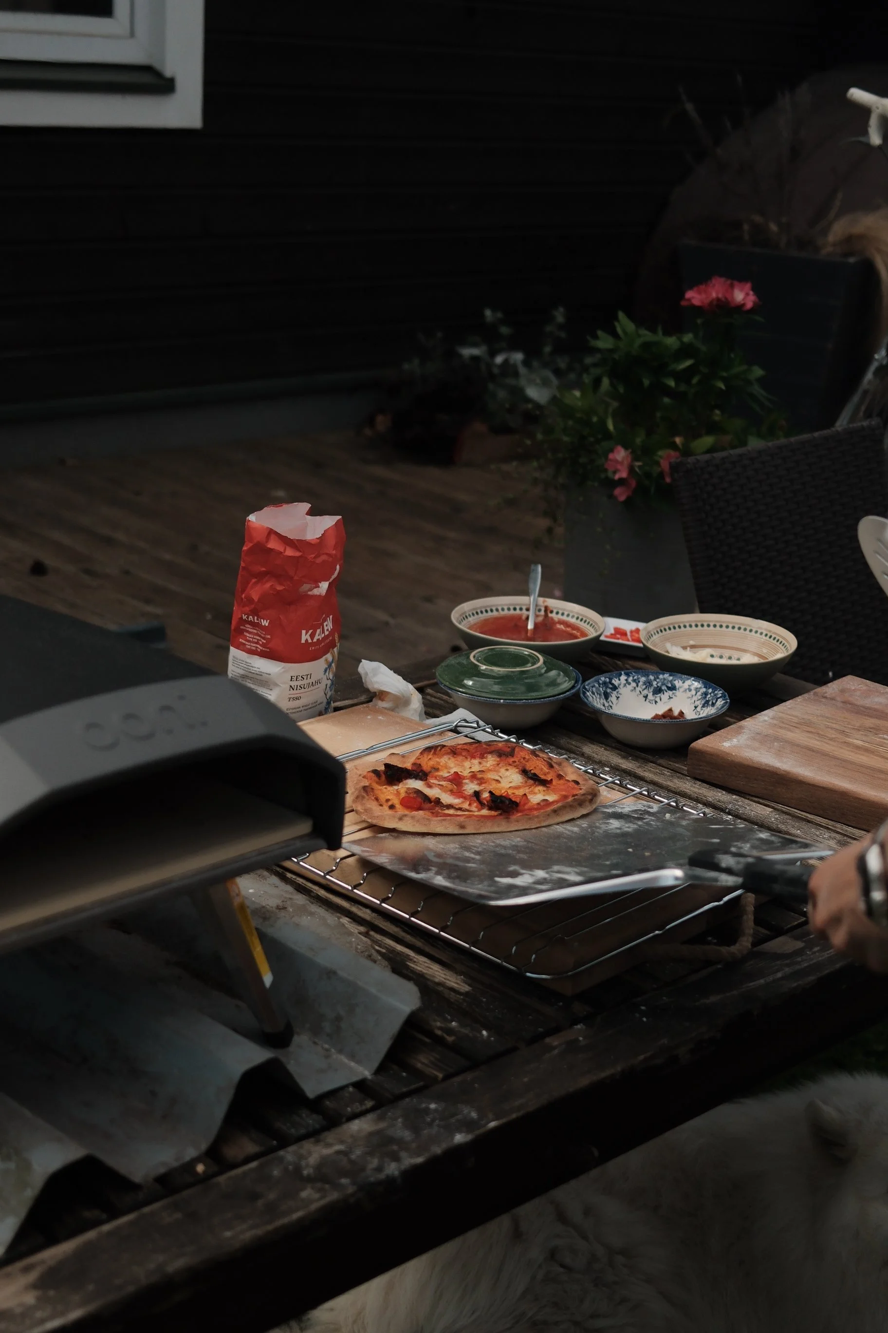 Scene of an outdoor dining area at dusk with a pizza on a wire rack, bowls of sauce, a bag of flour, and other kitchen items on a wooden table, with a flowerpot in the background.