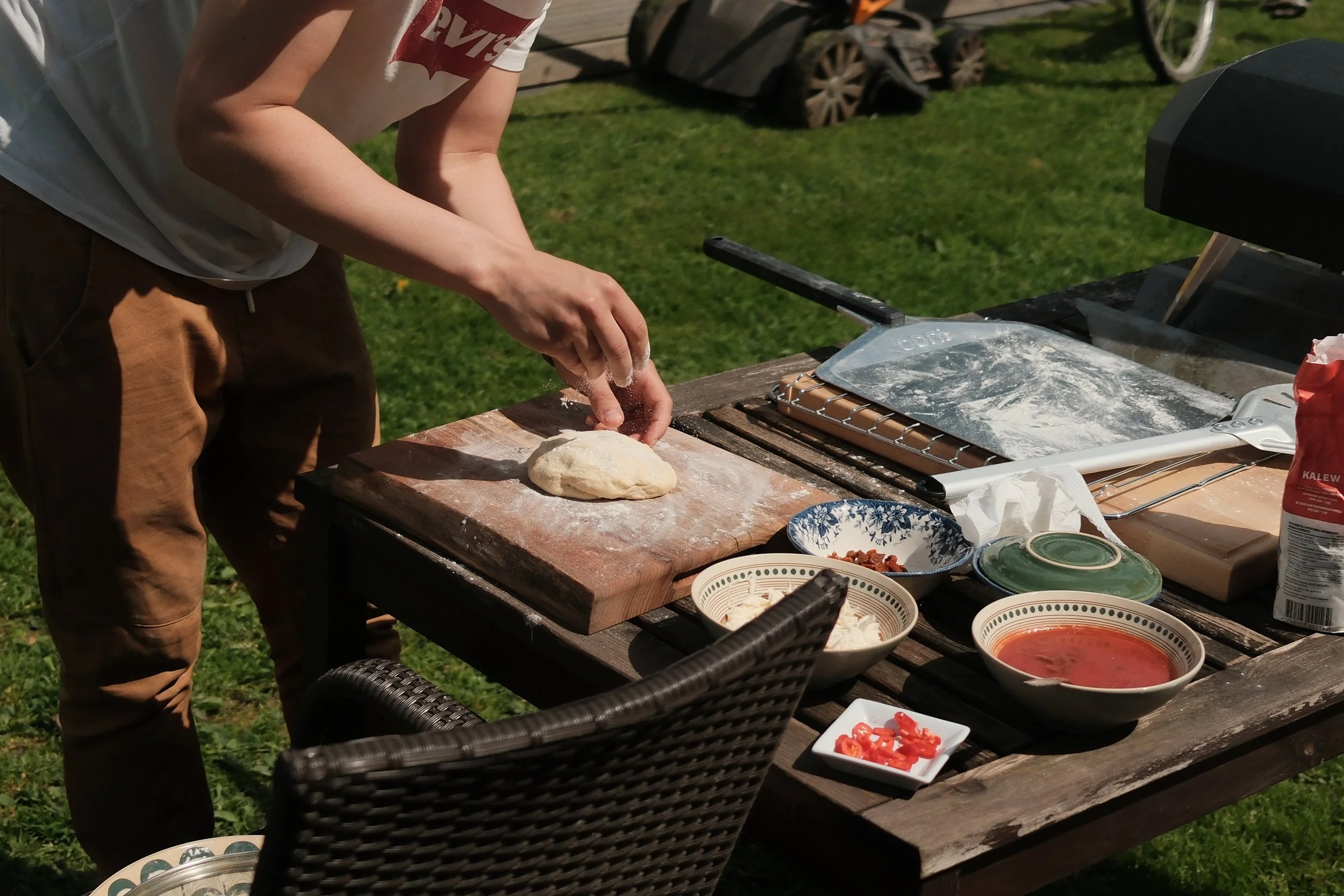 Close-up of a person kneading dough on a wooden outdoor table surrounded by bowls of ingredients and cooking utensils, with a grassy yard and garden in the background.