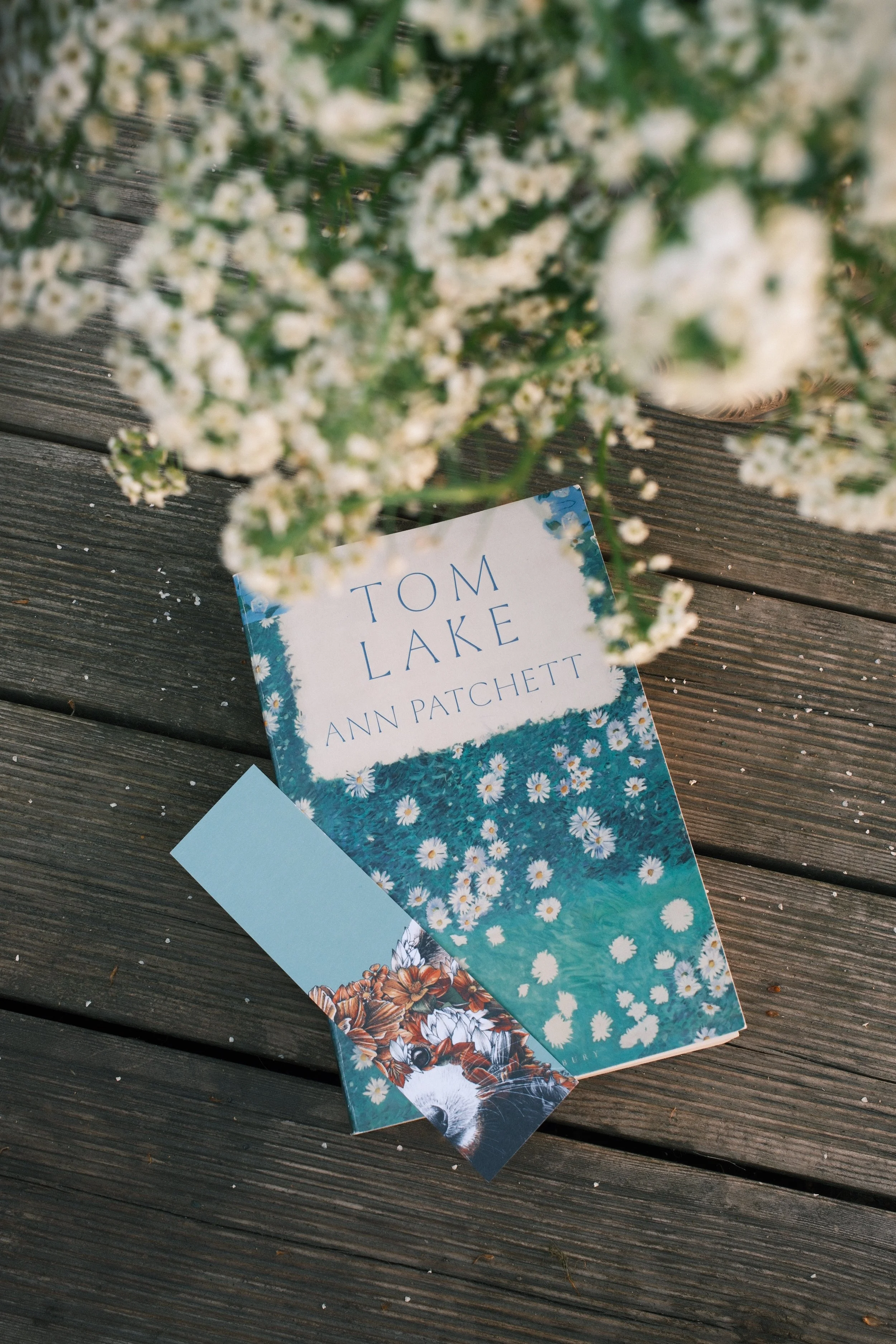 A wooden surface with a book titled 'Tom Lake' by Ann Patchett, partially covered by a bouquet of white flowers with green leaves.