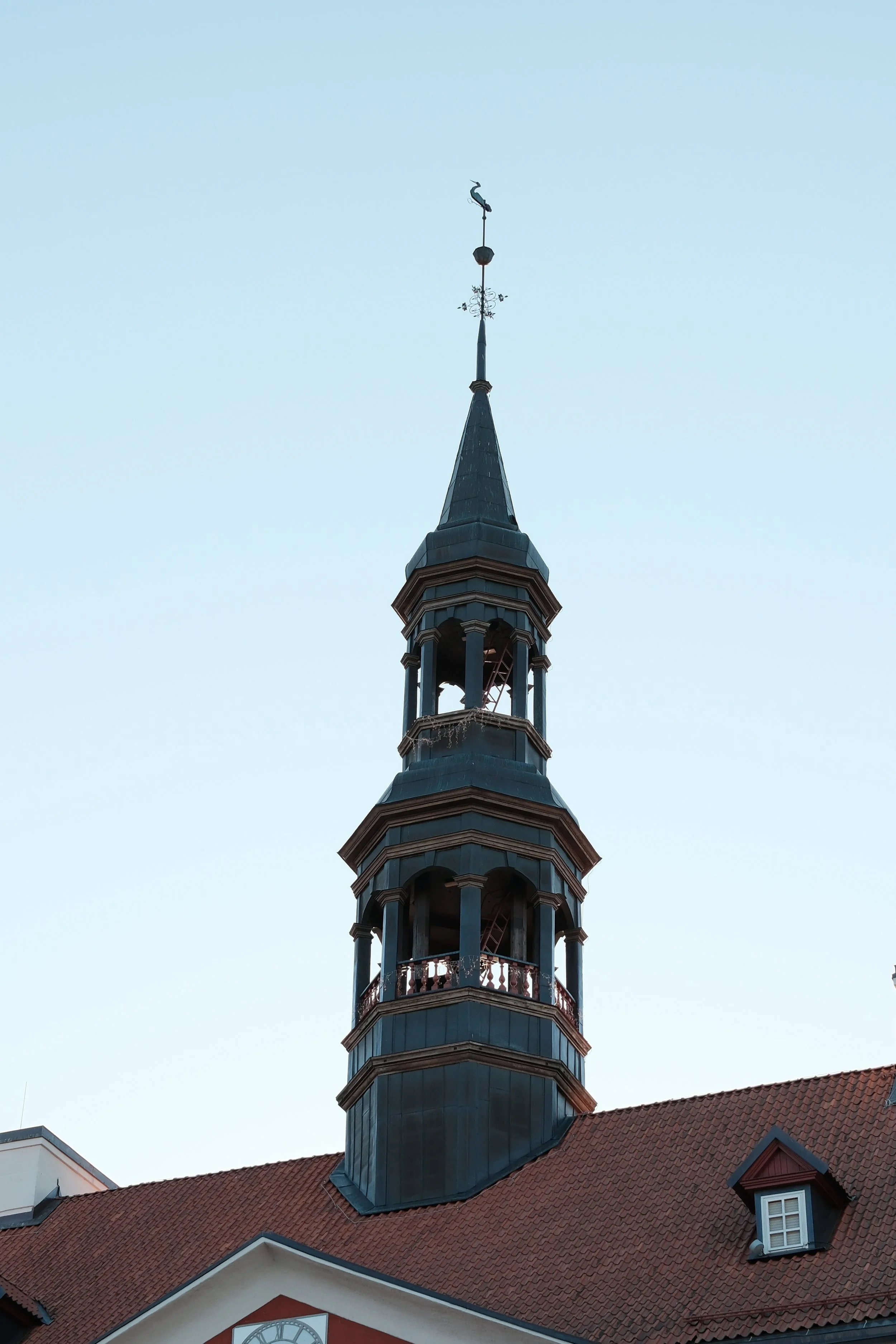 A tall church steeple with a weather vane in the shape of a heron at the top, attached to a building with a red tiled roof and dormer windows, against a clear blue sky.