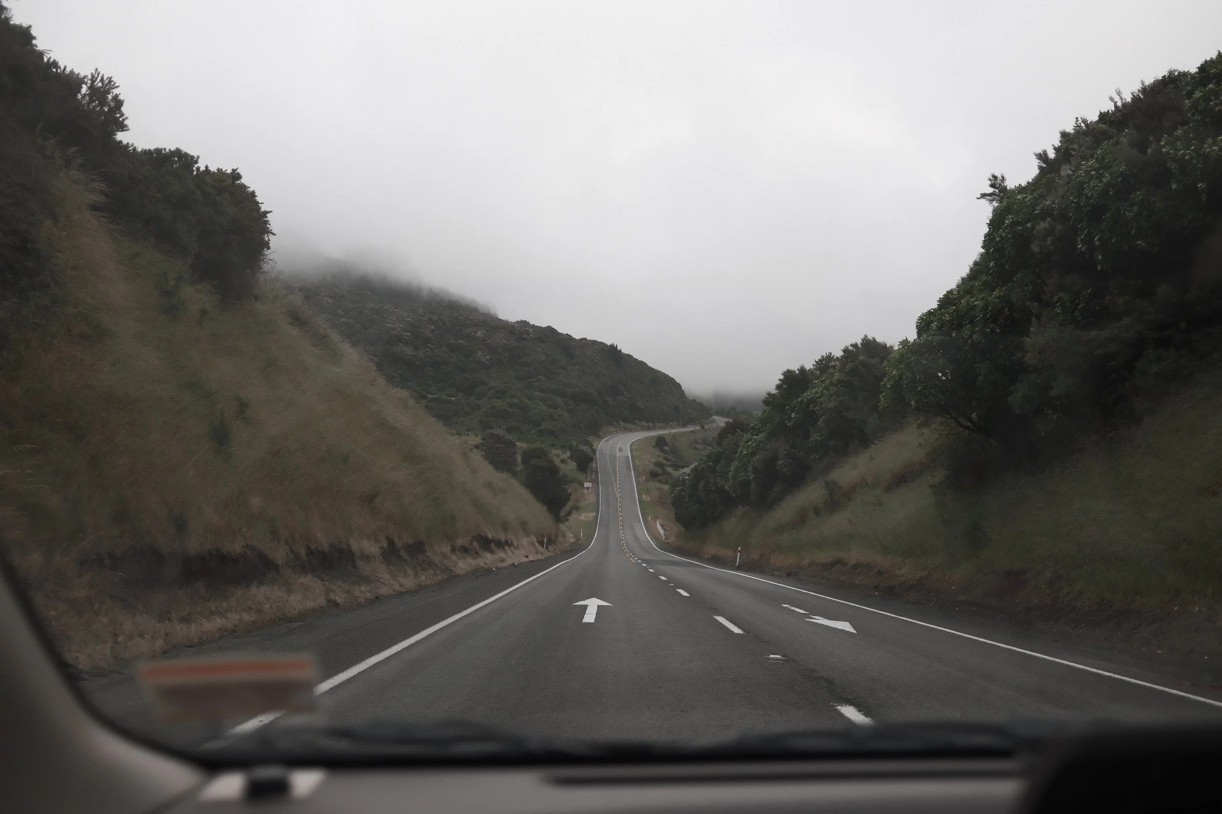 A winding mountain road viewed from inside a vehicle, with trees and hills on either side, and fog covering the distant hills.
