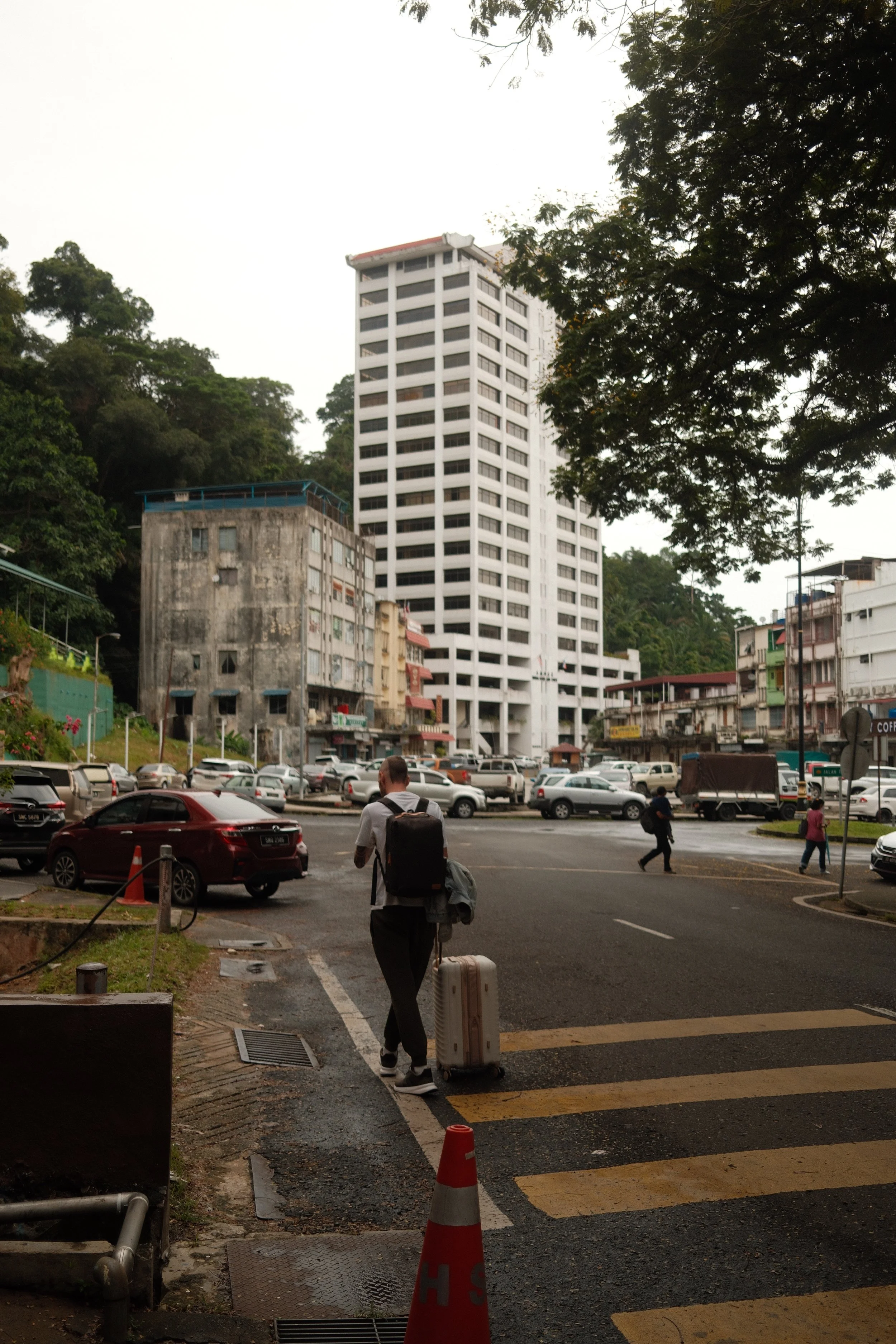 A man walking with a wheeled suitcase in a parking lot near a crosswalk, with a tall white building, smaller buildings, and a hillside covered with trees in the background.