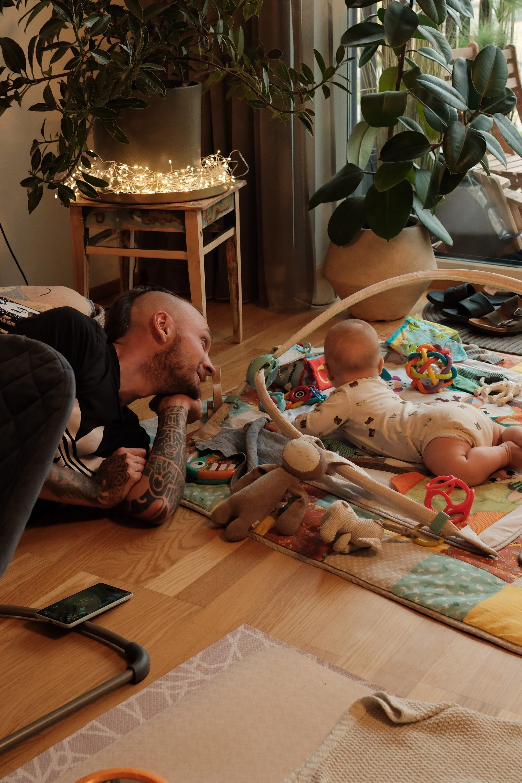 A man and a baby lying on a colorful play mat on the wooden floor inside a house. The man, with tattoos and a beard, is resting his head on his hand and smiling, looking at the baby. The baby, dressed in beige with brown pattern, is playing with colo