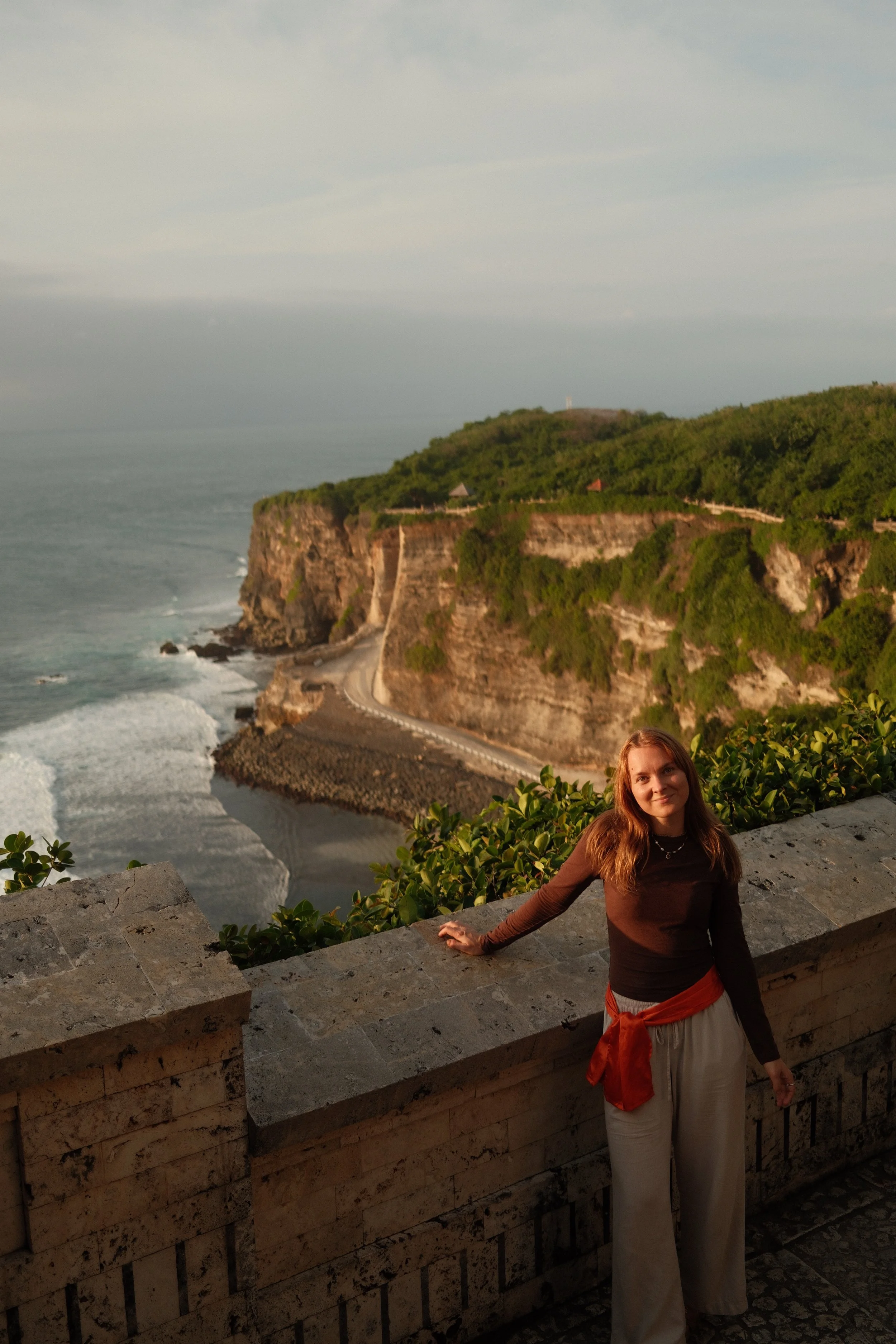 A woman with long hair wearing a brown top and wide beige pants stands leaning against a stone railing overlooking a coastal landscape with cliffs, greenery, and the ocean, during sunset.