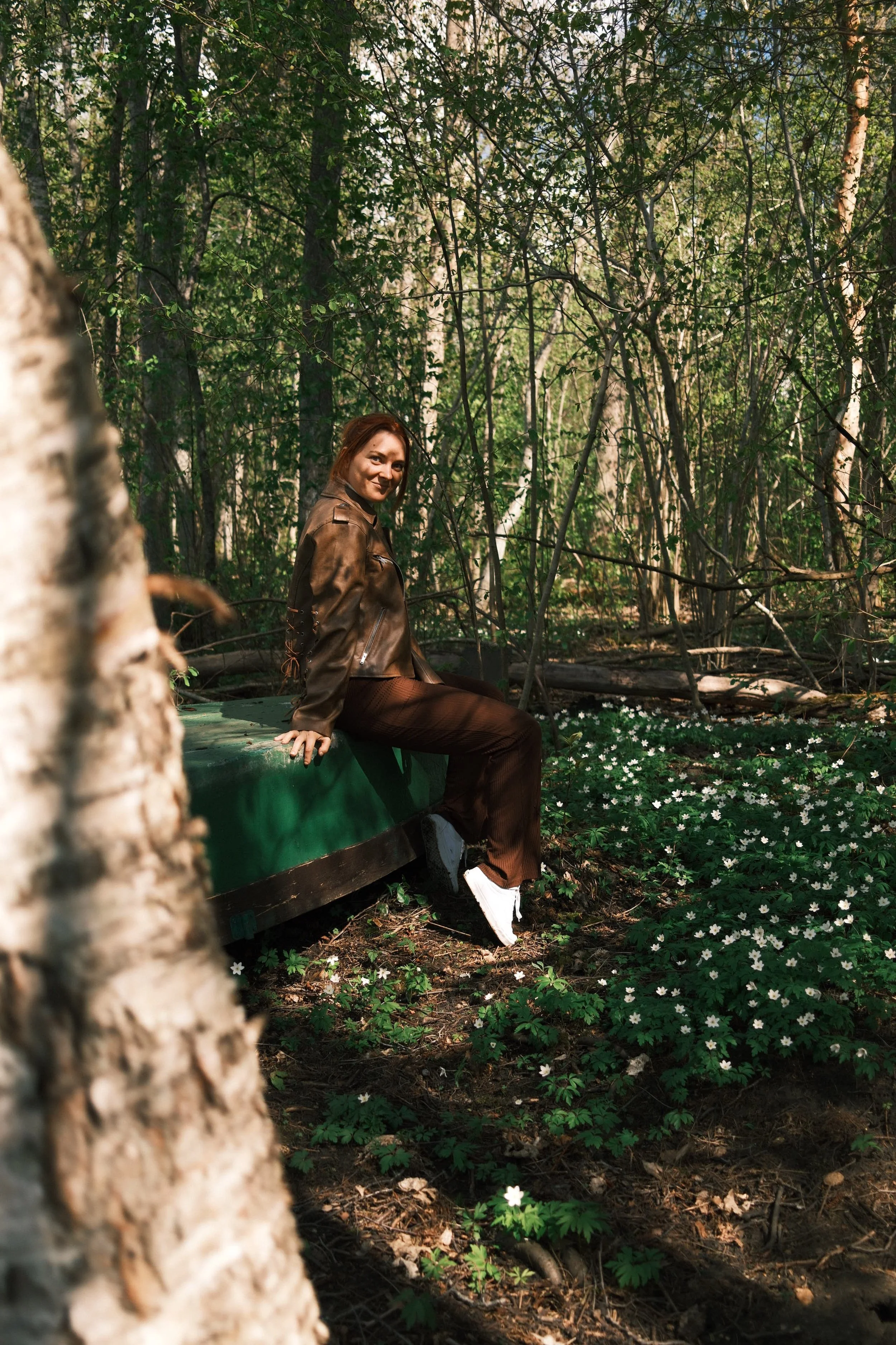 A woman sitting on a green boat in a forest, surrounded by trees and white flowers on the ground, smiling at the camera.