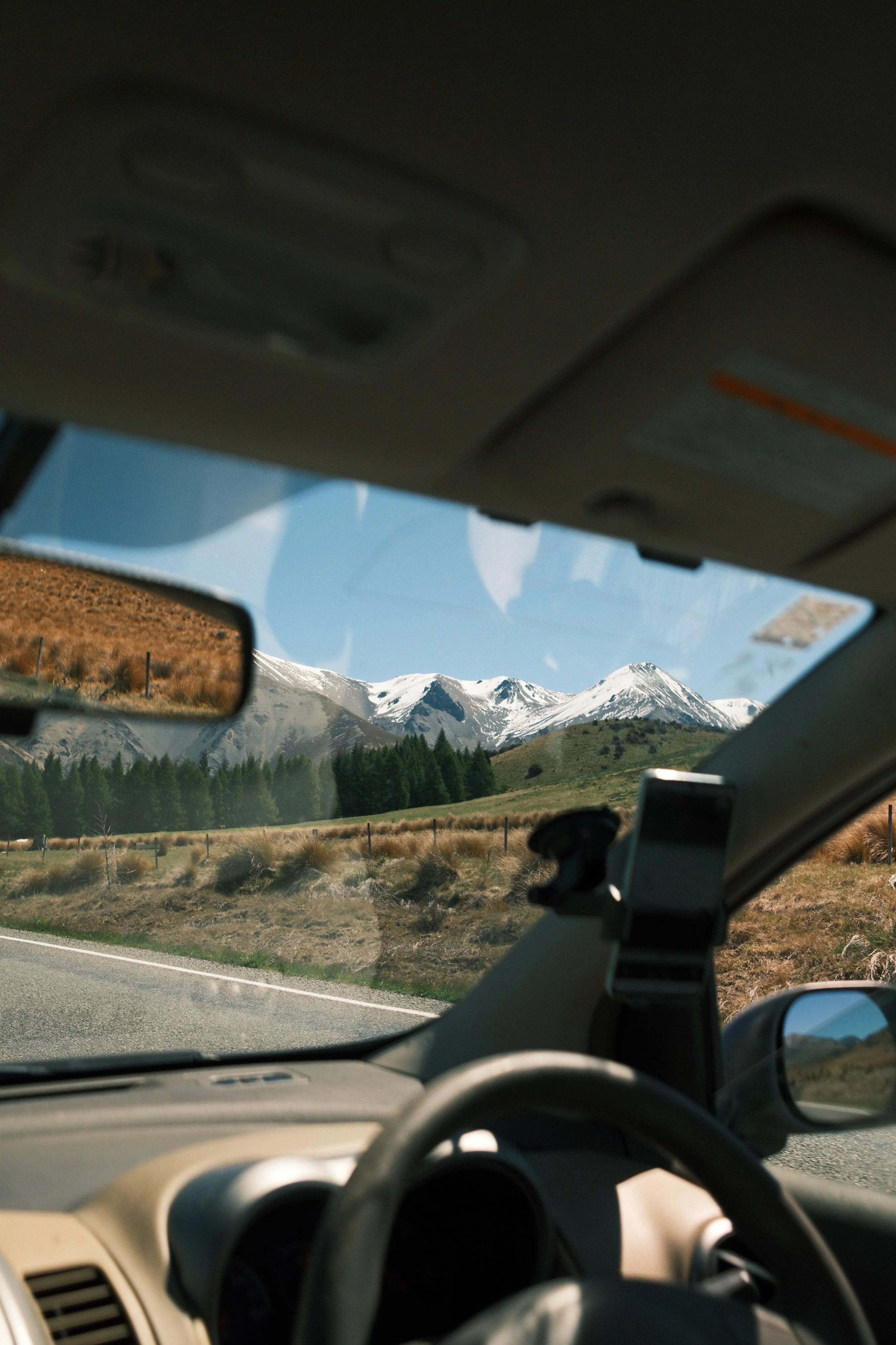 Photo taken from inside a car showing snow-capped mountains through the windshield, with a side mirror reflecting a mountain scene, road, and landscape.