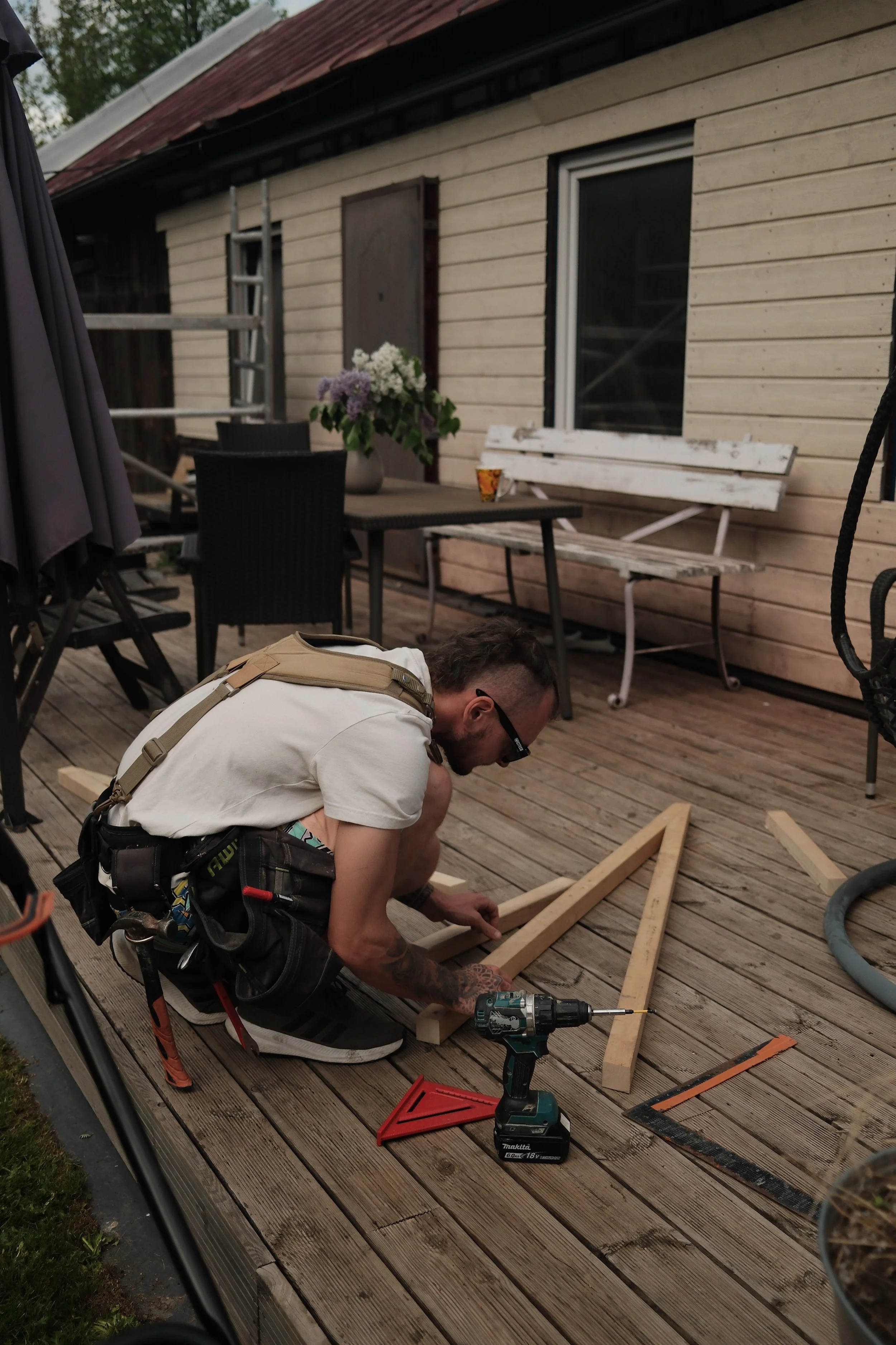 A man wearing glasses, a white shirt, and a tool belt is working on assembling a wooden frame on a wooden deck outside a house. The house in the background has beige siding, a window, and a white bench with chipped paint. There is also a vase with pu