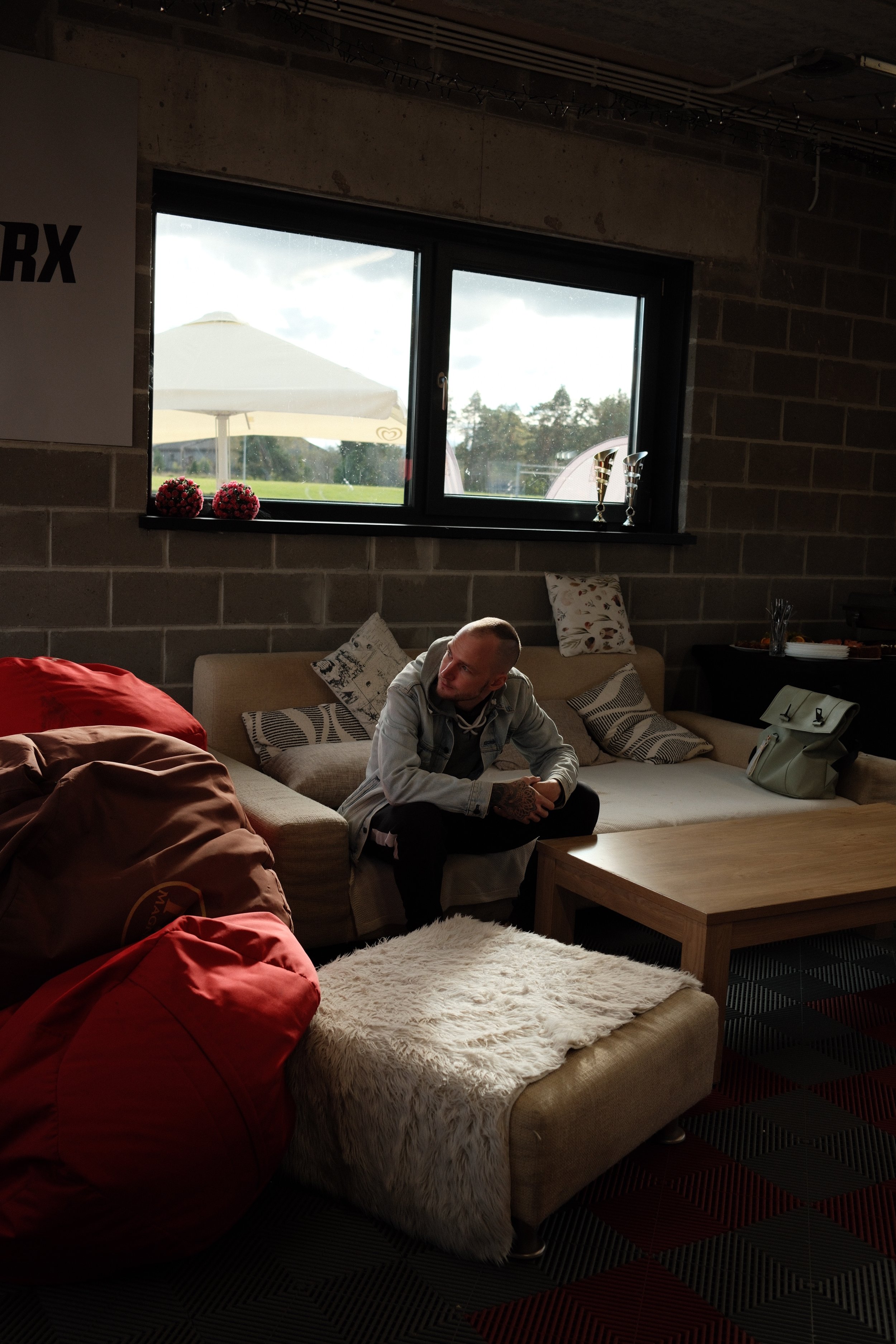 A man sitting on a beige couch with multiple pillows, some with patterned covers, in a room with a brick wall. There is a white fluffy blanket on the couch, a wooden coffee table. Outside the window, a large umbrella and a grassy area with trees are 