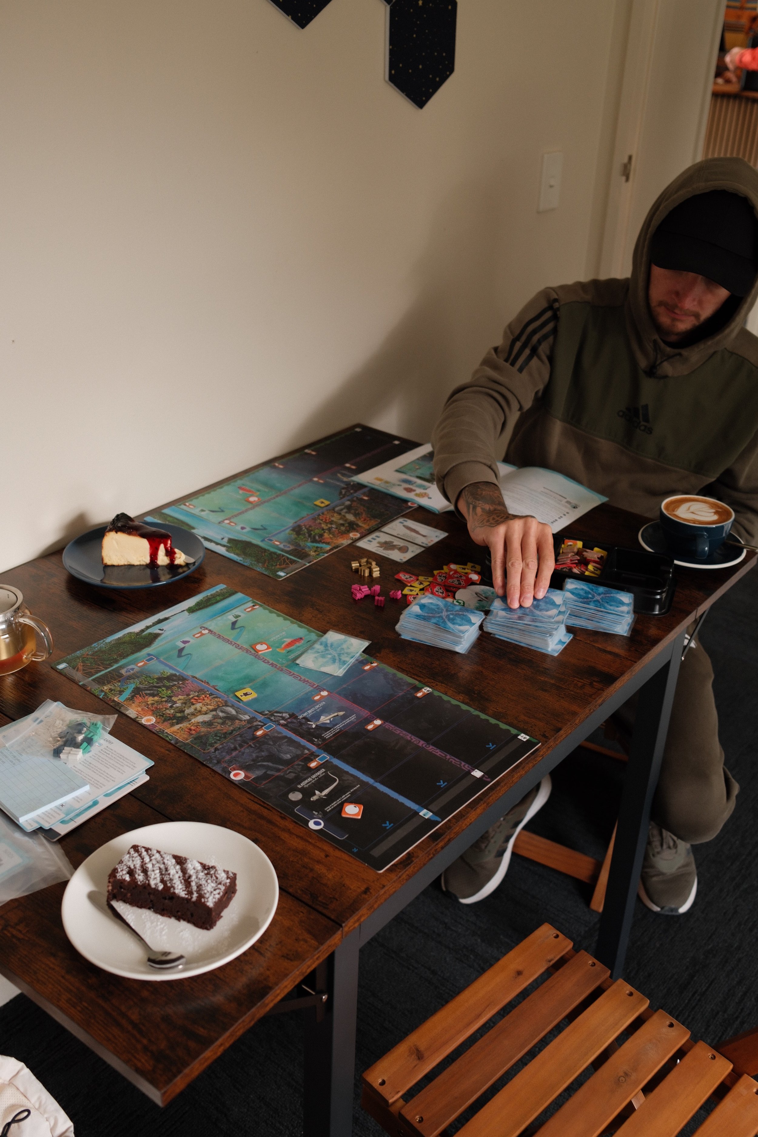 A man wearing a hoodie and black cap sitting at a wooden table with board games and a cup of coffee. There is a slice of cake on a black plate, a slice of cake on a white plate, and a rectangular chocolate cake in the foreground.
