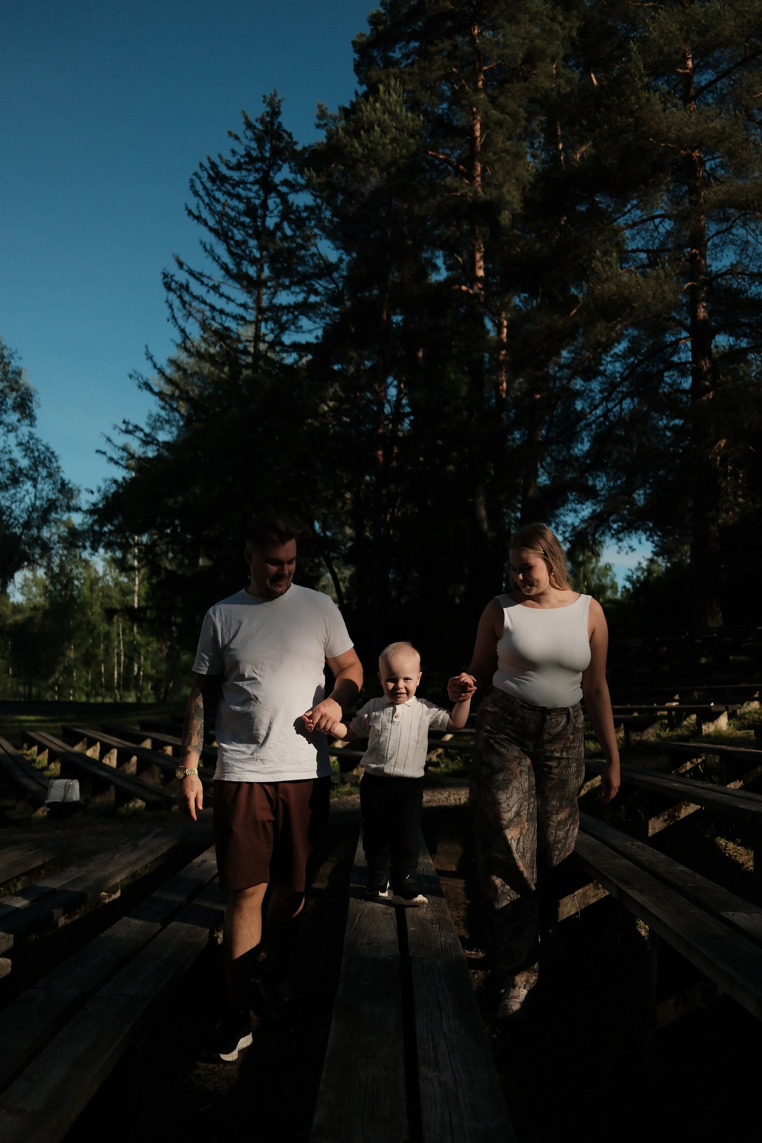 A family of four walking together outdoors on a wooden bridge in the evening, surrounded by trees and a blue sky.