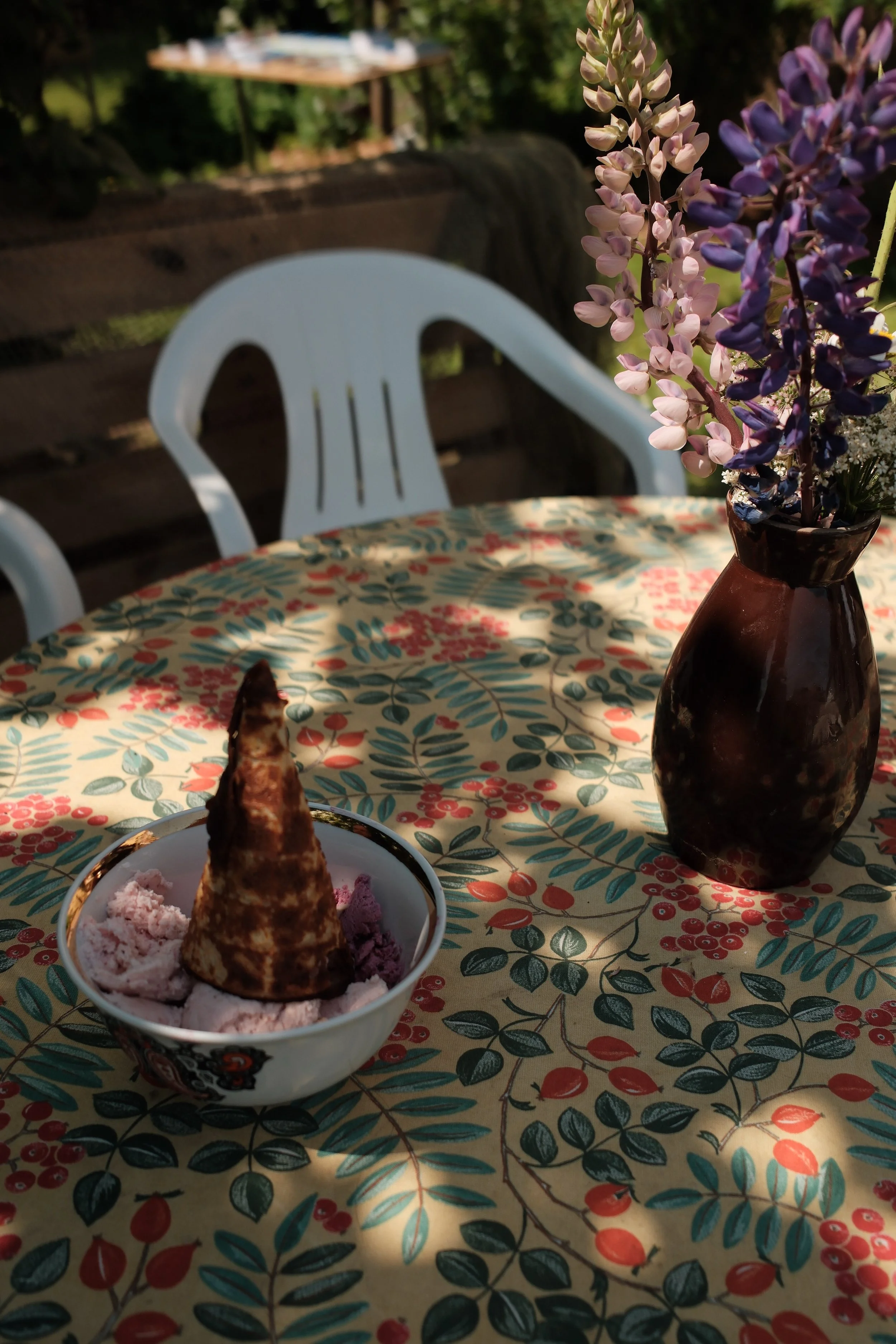 Ice cream in a bowl topped with a cone, on a table with a floral tablecloth, with a vase of purple and white flowers nearby. In the background, there are outdoor tables and chairs.
