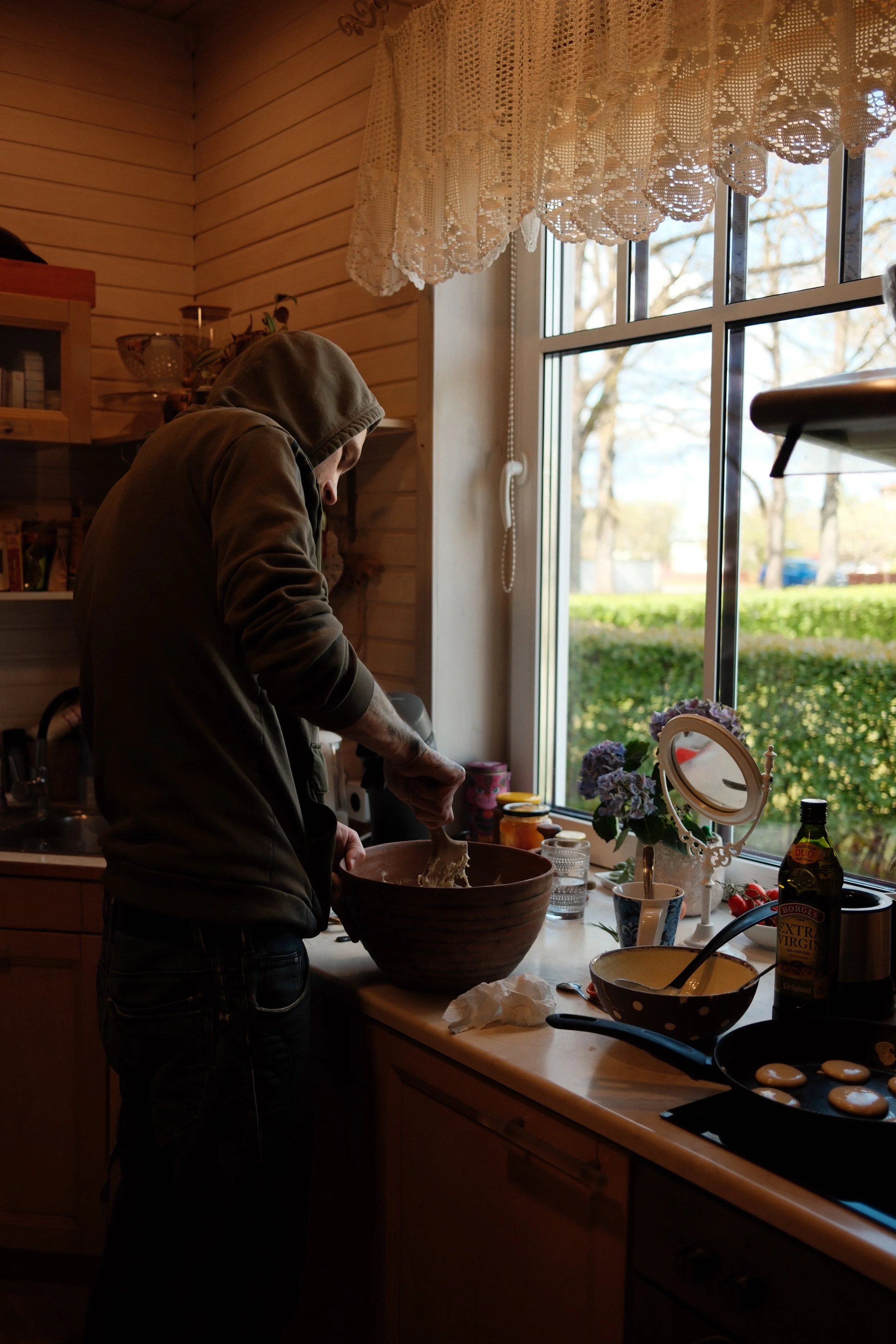 A person in a hoodie preparing food in a cozy kitchen with natural light coming through a large window. The kitchen decor includes a lace curtain, flowers, and various kitchen items on the counter including a bowl of eggs, a bottle of extra virgin ol