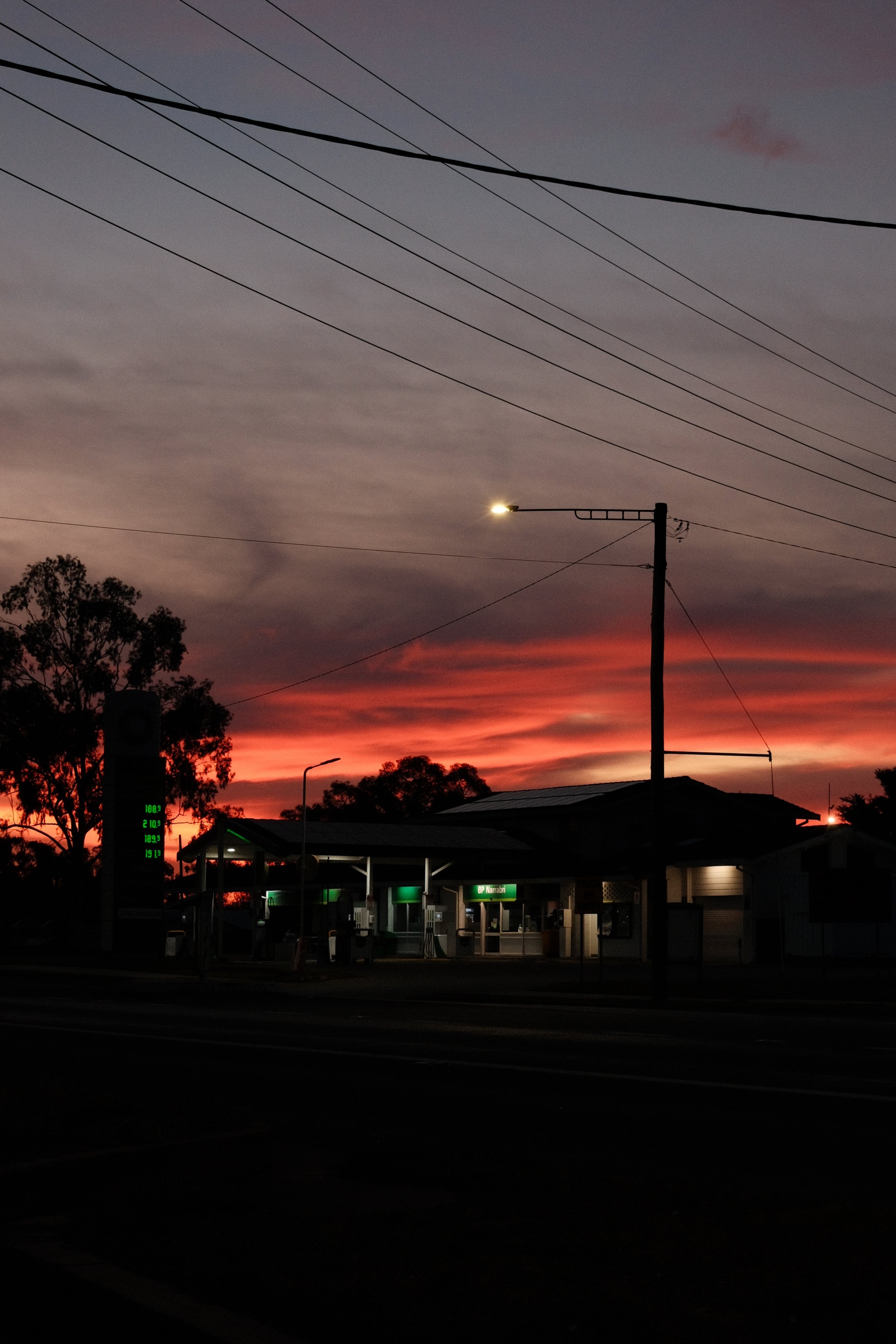 A sunset sky with clouds and a gradient of colors from dark purple to orange, with power lines crossing the sky and a gas station with illuminated signs in the foreground.