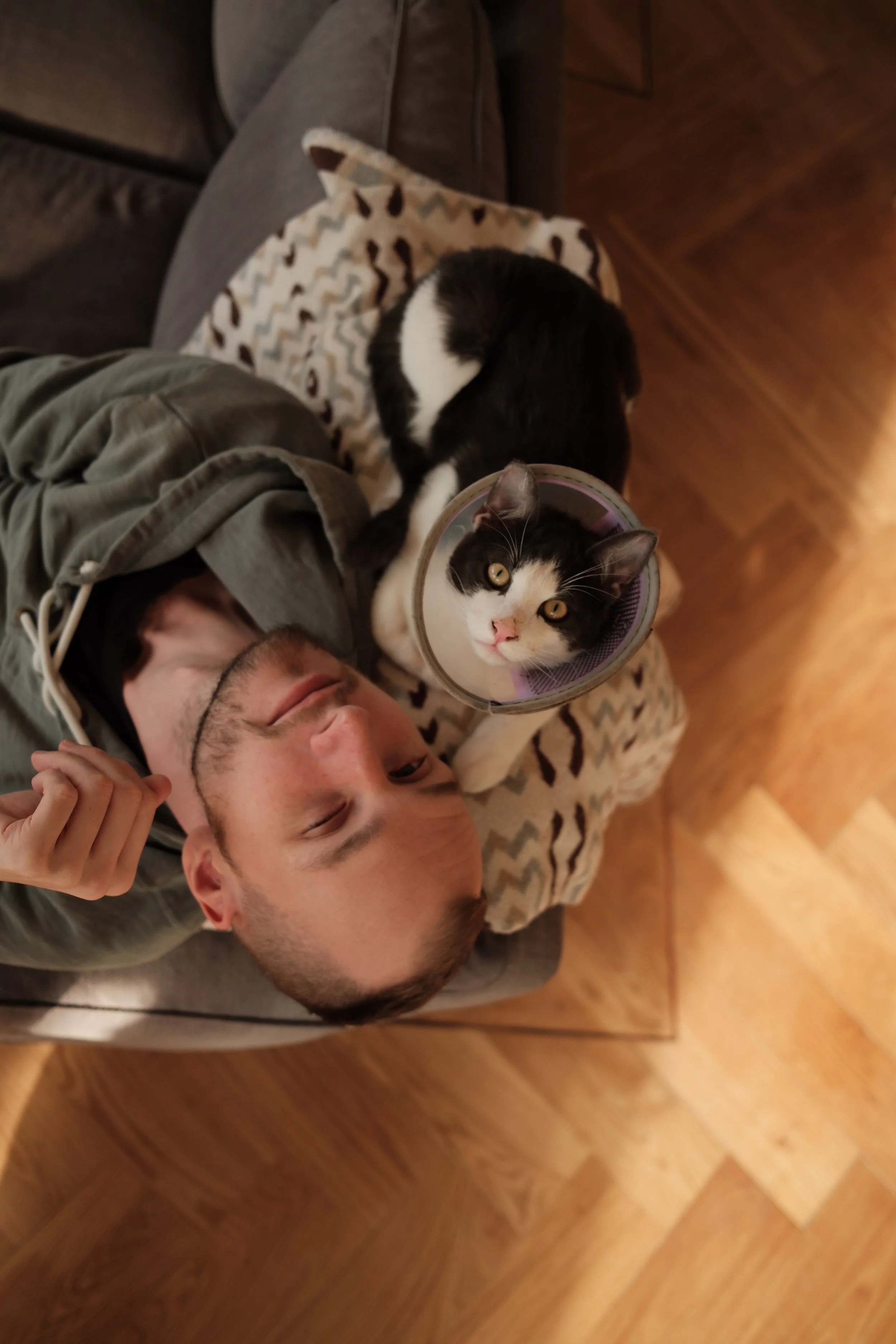 A man lying on a wooden floor with two black and white cats, one cat on the man's shoulder and the other sleeping on a patterned blanket.
