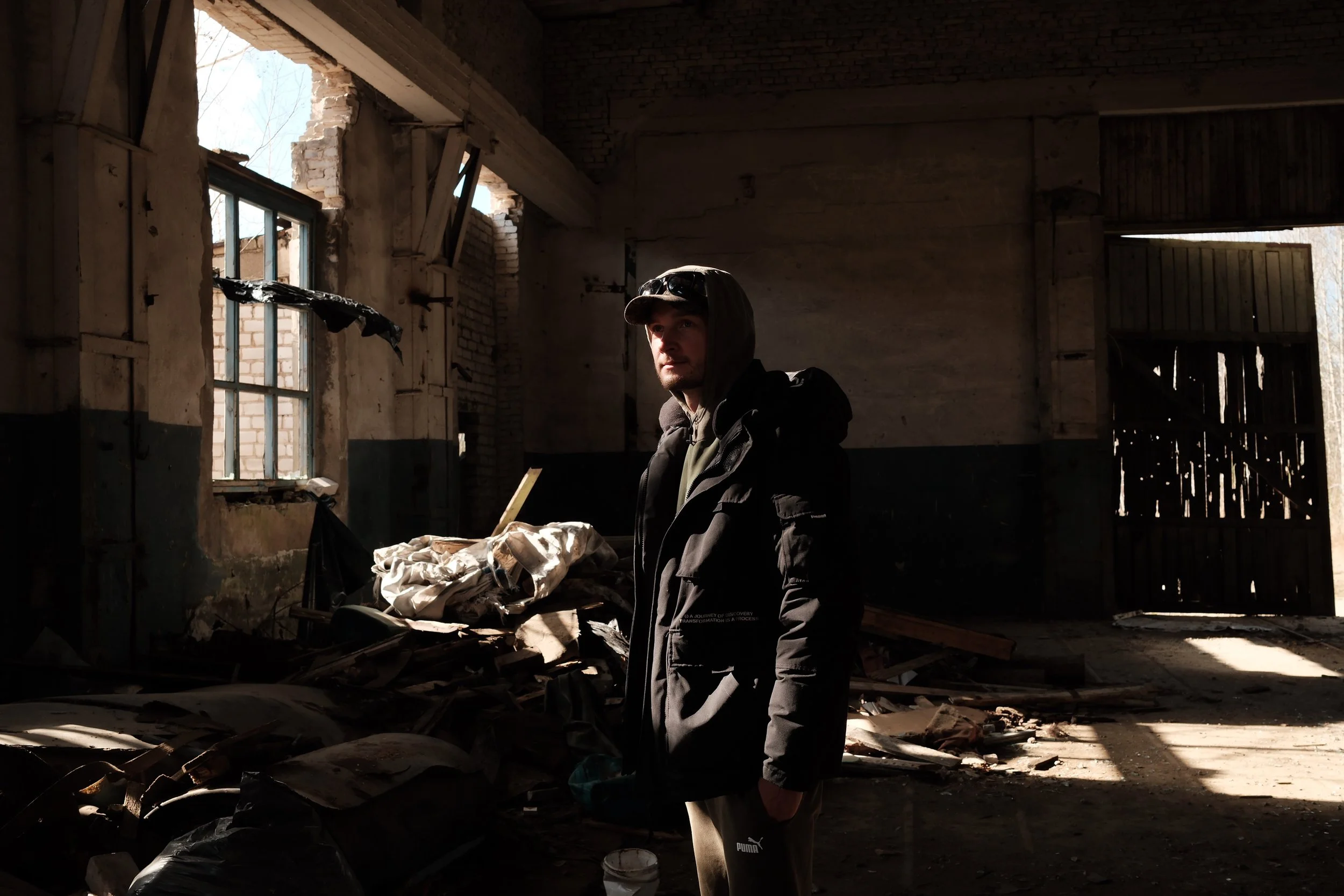 A man standing inside an abandoned, dilapidated building with broken windows and scattered debris, illuminated by natural light coming through openings in the walls and roof.