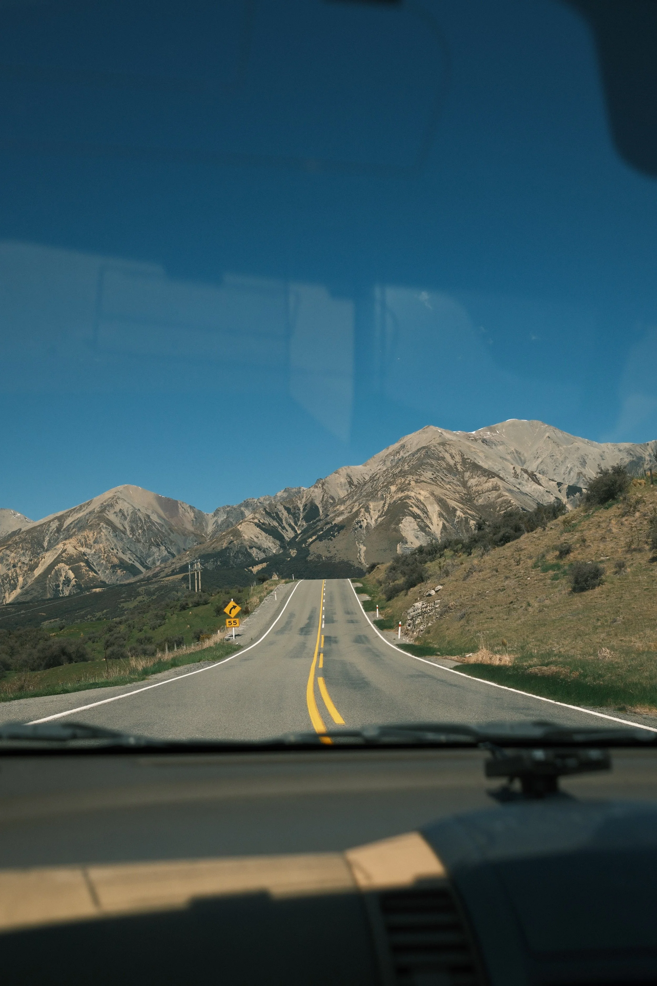 View of a winding mountain road taken from inside a vehicle, with mountains in the background and clear blue sky.