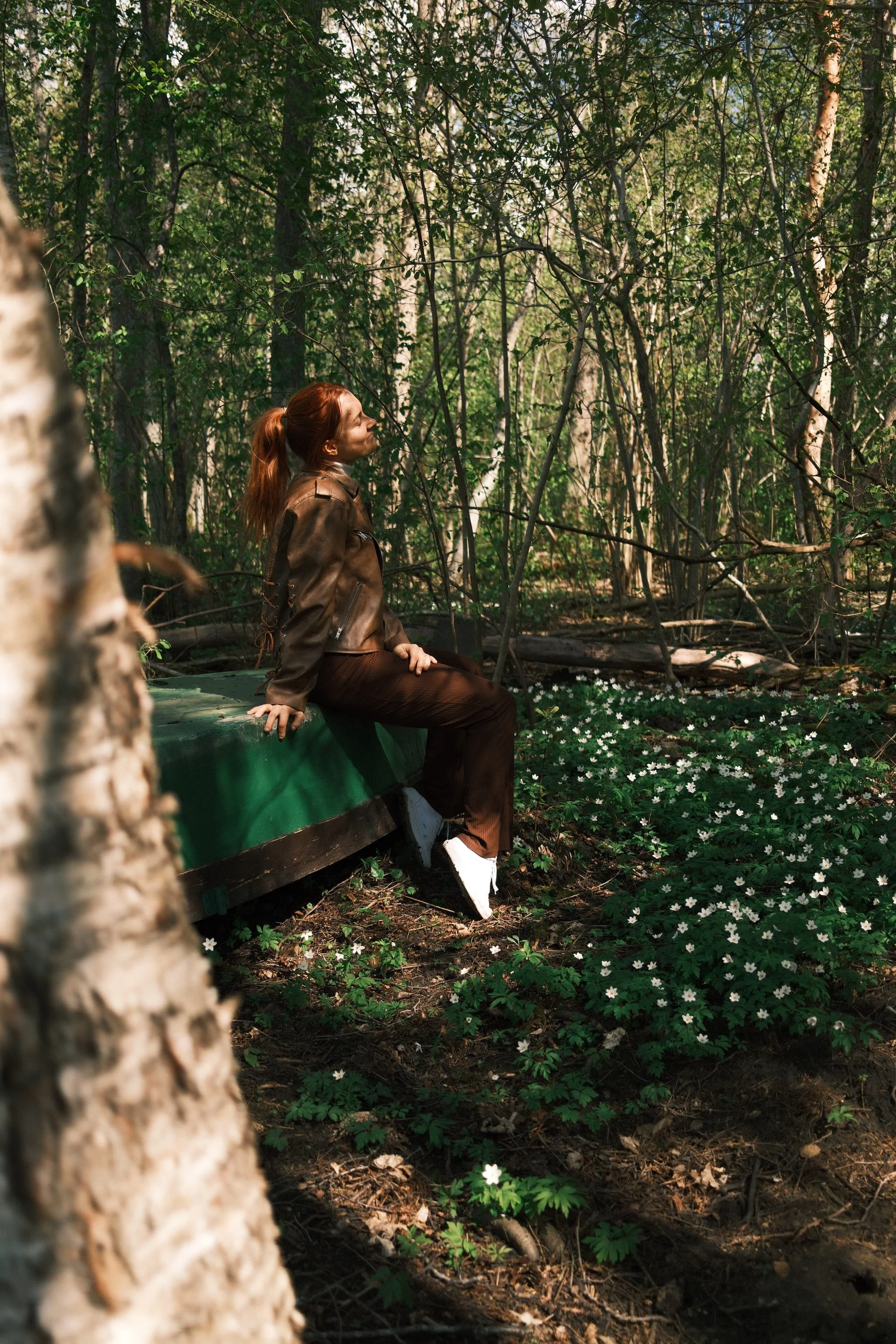 A woman with red hair sitting on a green boat in a forested area, surrounded by trees and white flowers.